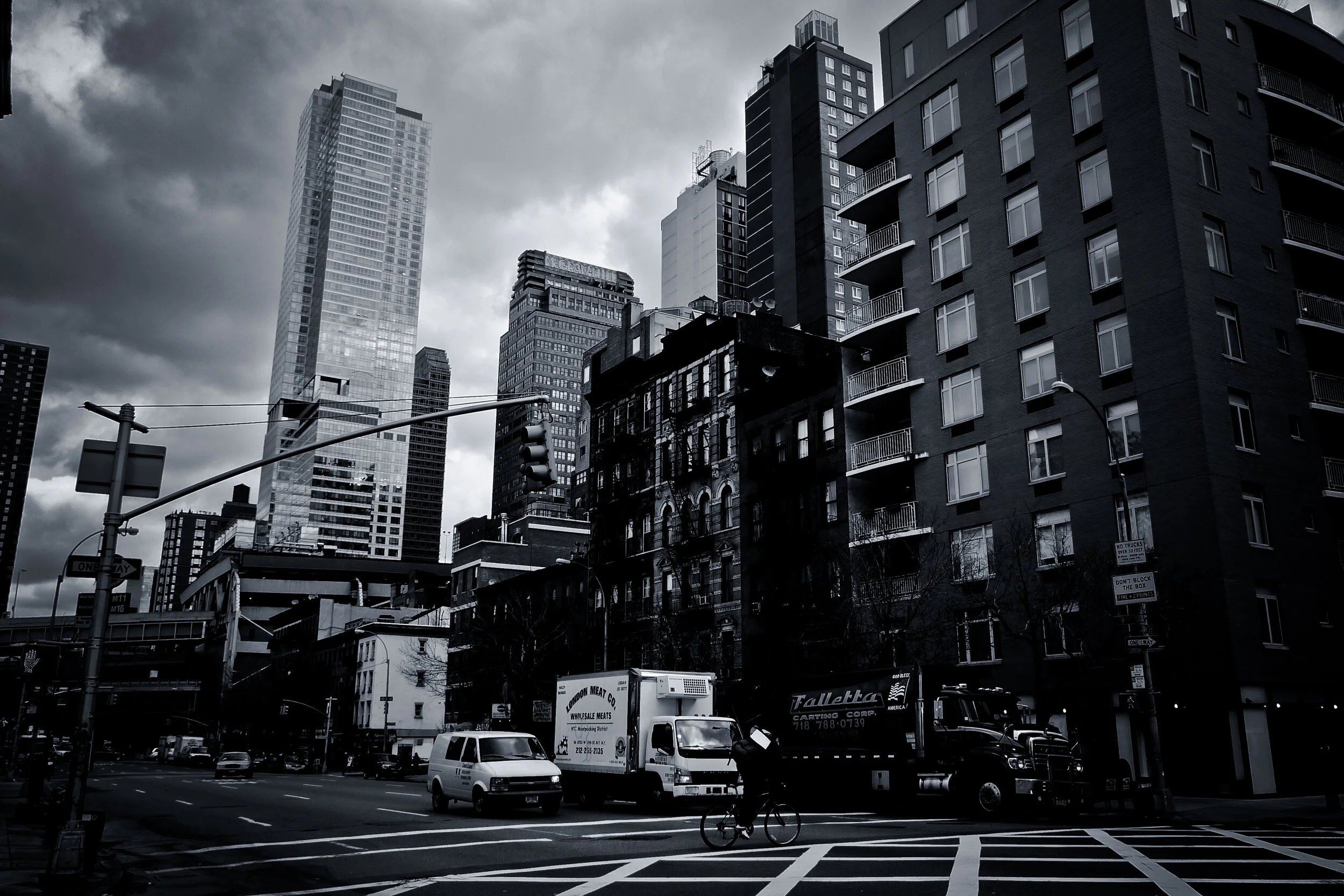 Black and white photo of an urban cityscape with tall buildings, cloudy sky, traffic signals, and vehicles on the street.