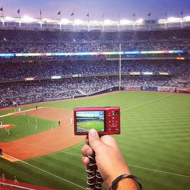 A person taking a photo of a baseball game at a stadium with a digital camera, baseball field and players visible in the background, and spectator stands filled with audience under a cloudy sky.