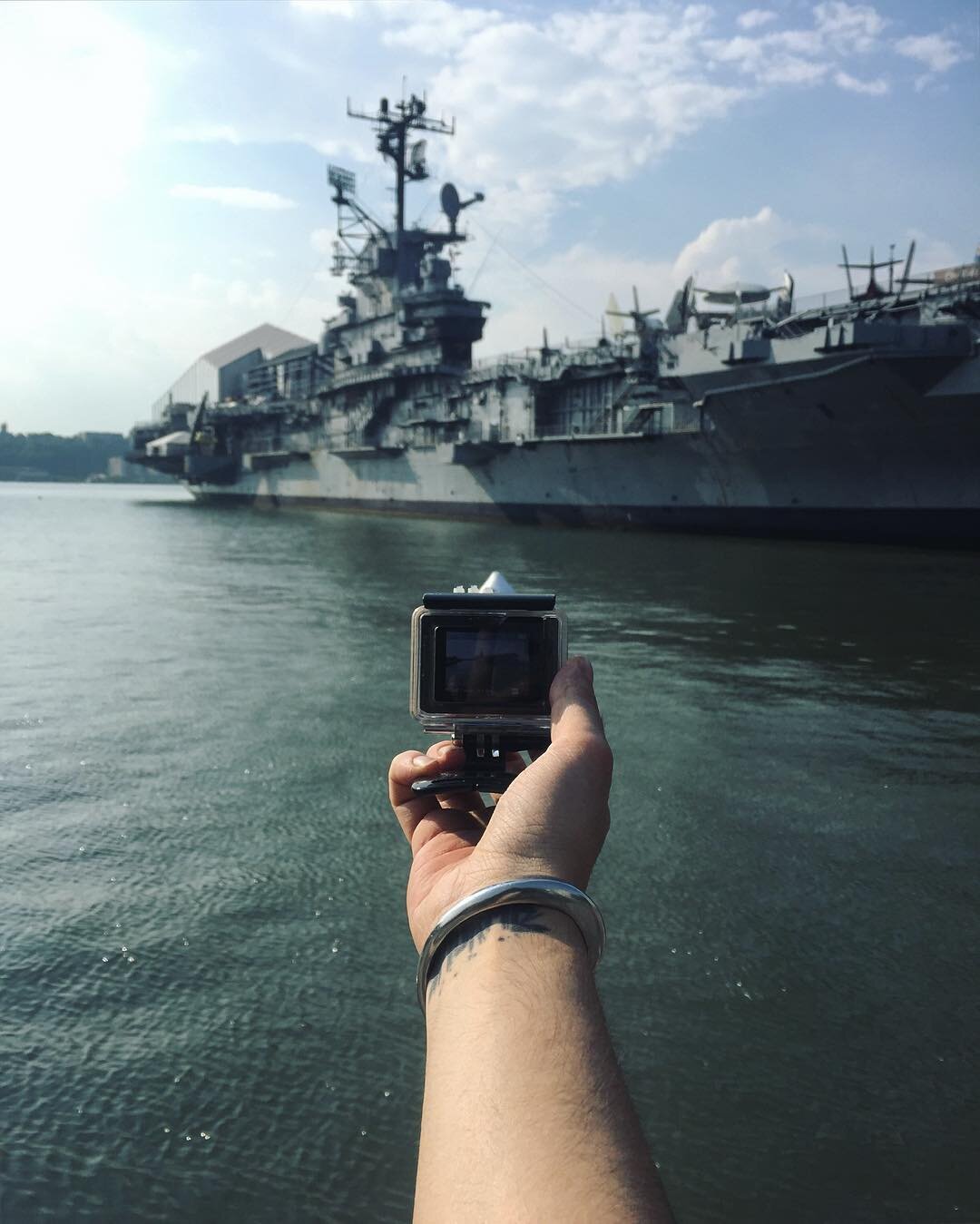Person holding a camera taking a photo of a large gray military battleship docked on the water at a harbor, with a partly cloudy sky.