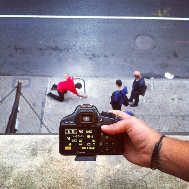 Photograph taken from above showing a person holding a Canon camera aiming down at a sidewalk. On the sidewalk, a man in a red shirt is kneeling by a phone booth, and two other men, one carrying a backpack, are walking by.