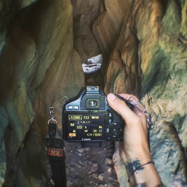 A person holding a digital camera takes a photo inside a narrow, rocky crevice or tunnel with textured, multi-colored rock walls.