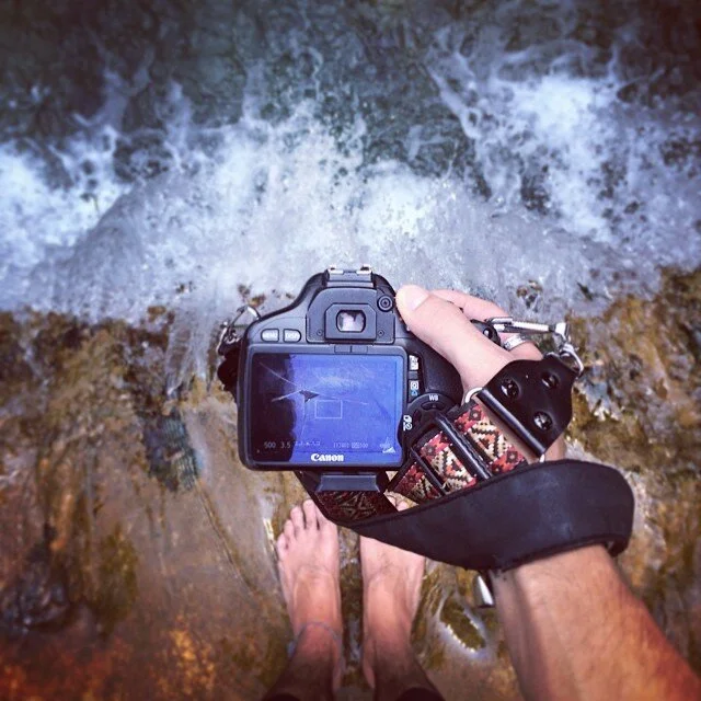 Person standing in a shallow creek taking a photograph of the water with a Canon camera, visible bare feet, and rocky bed.
