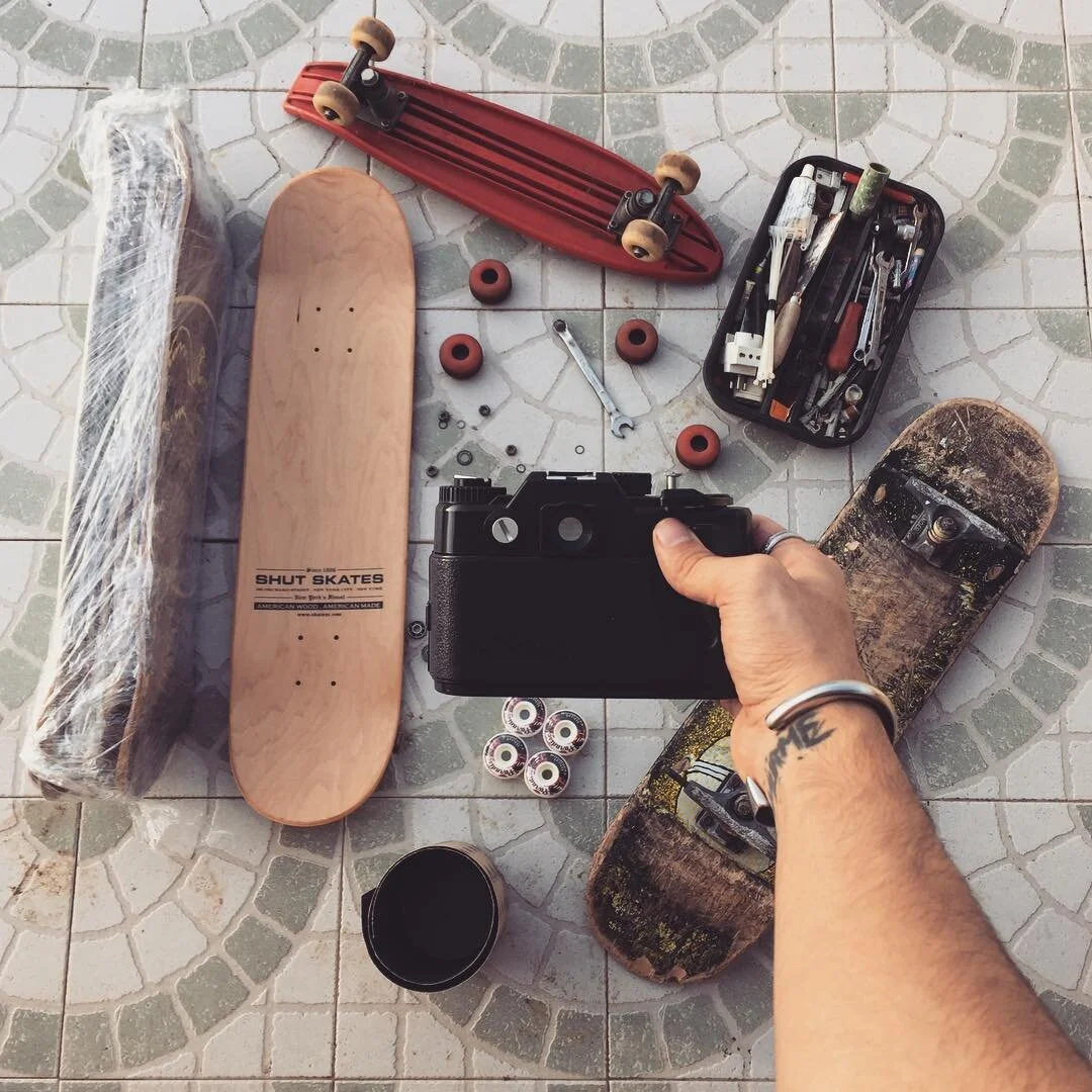 Various skateboards, skate tools, wheels, and skateboard parts laid out on a tiled floor, with a person holding a camera in the foreground.