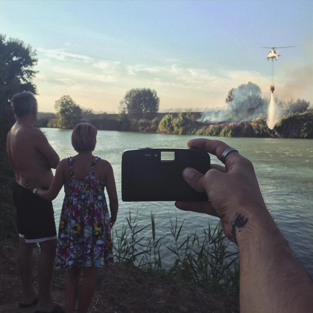 People by a river watching a helicopter spraying water on a fire in the distance.