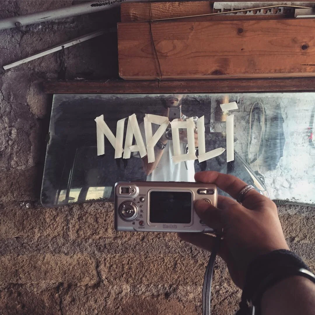 Person taking a photo of a mirror with a compact camera, which reflects the word "NAPLIL" spelled out with tape. The mirror is mounted on a stone wall above a wooden surface.