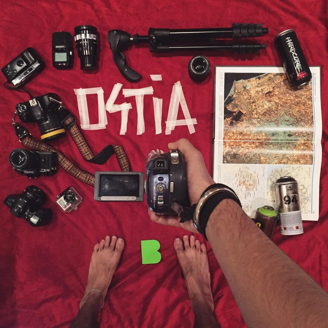 A red cloth spread with various photography and outdoor gear, including camera equipment, a map, and soda cans, with the word 'ACTION' spelled out with tape and the person's toes visible in the foreground.