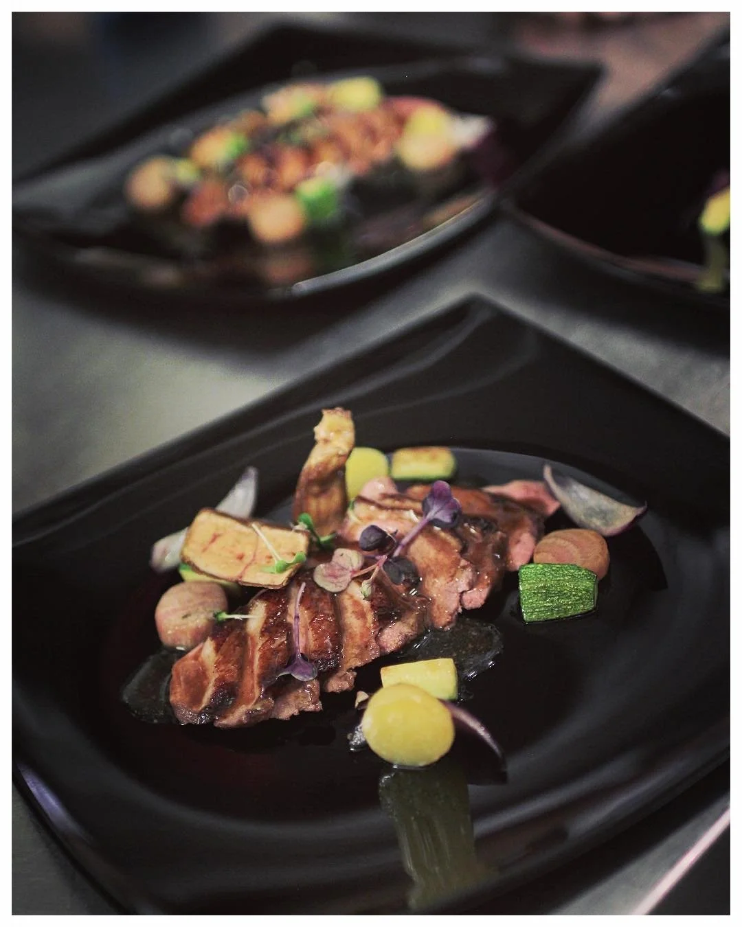 A plated dish featuring grilled meat garnished with various vegetables and microgreens, presented on a black plate.