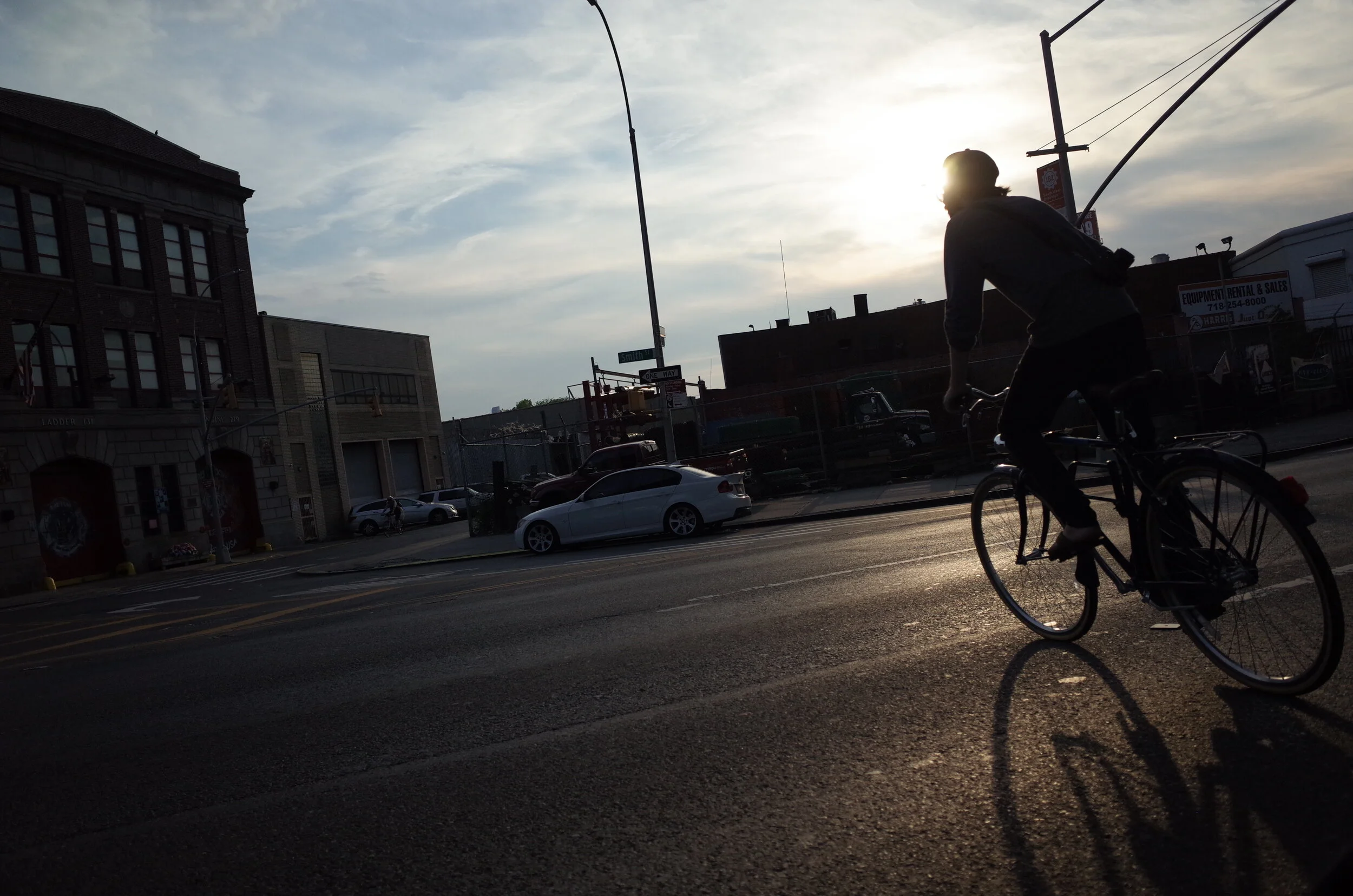 A person riding a bicycle on a city street during sunset, with cars parked along the side and buildings in the background.