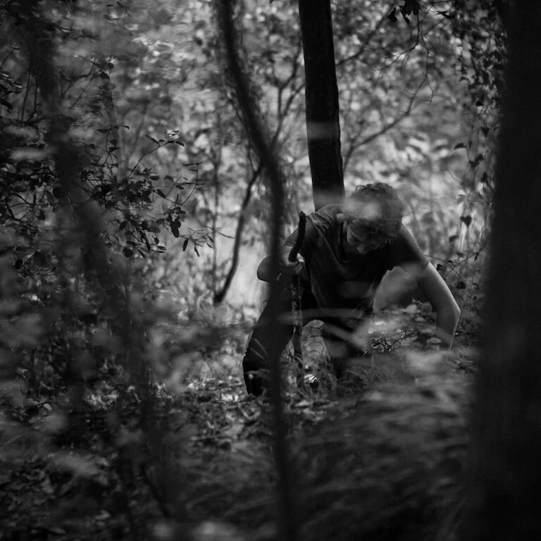 A person wearing glasses and a dark shirt, carrying a backpack, is climbing over a fallen tree in a dense forest with tall trees and thick foliage, in grayscale.