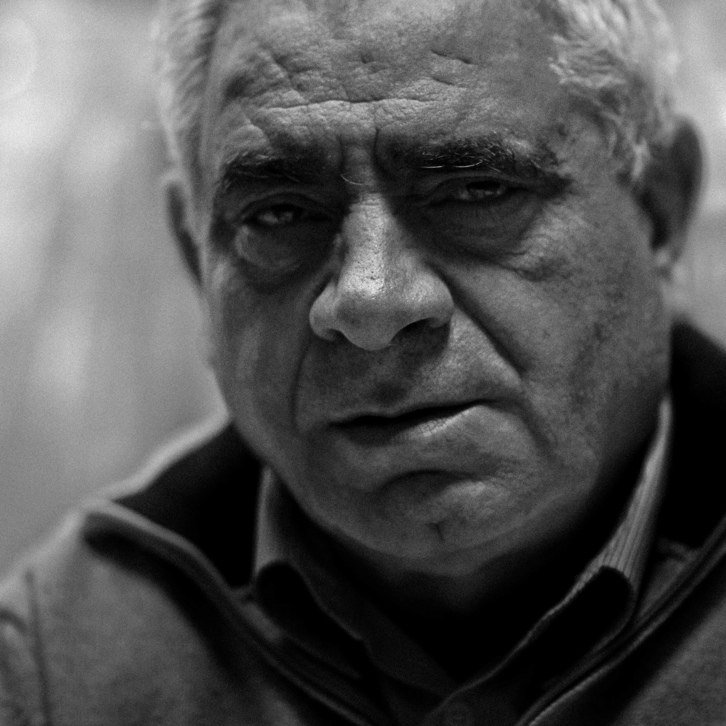 A close-up black and white photograph of an elderly man's face, focusing on his closed and partially open eyes, wrinkles, and textured skin.