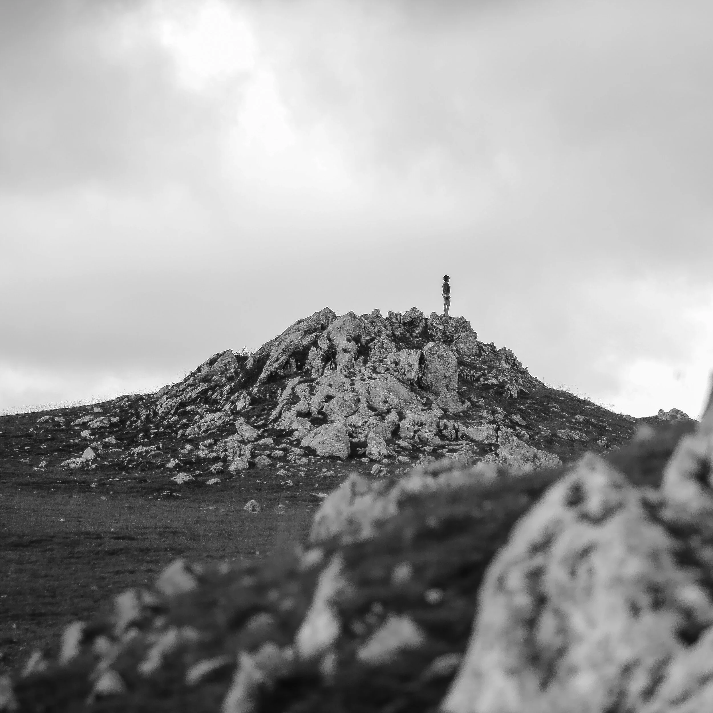 A person standing on top of a rocky mountain ridge, looking out at a cloudy sky in black and white.