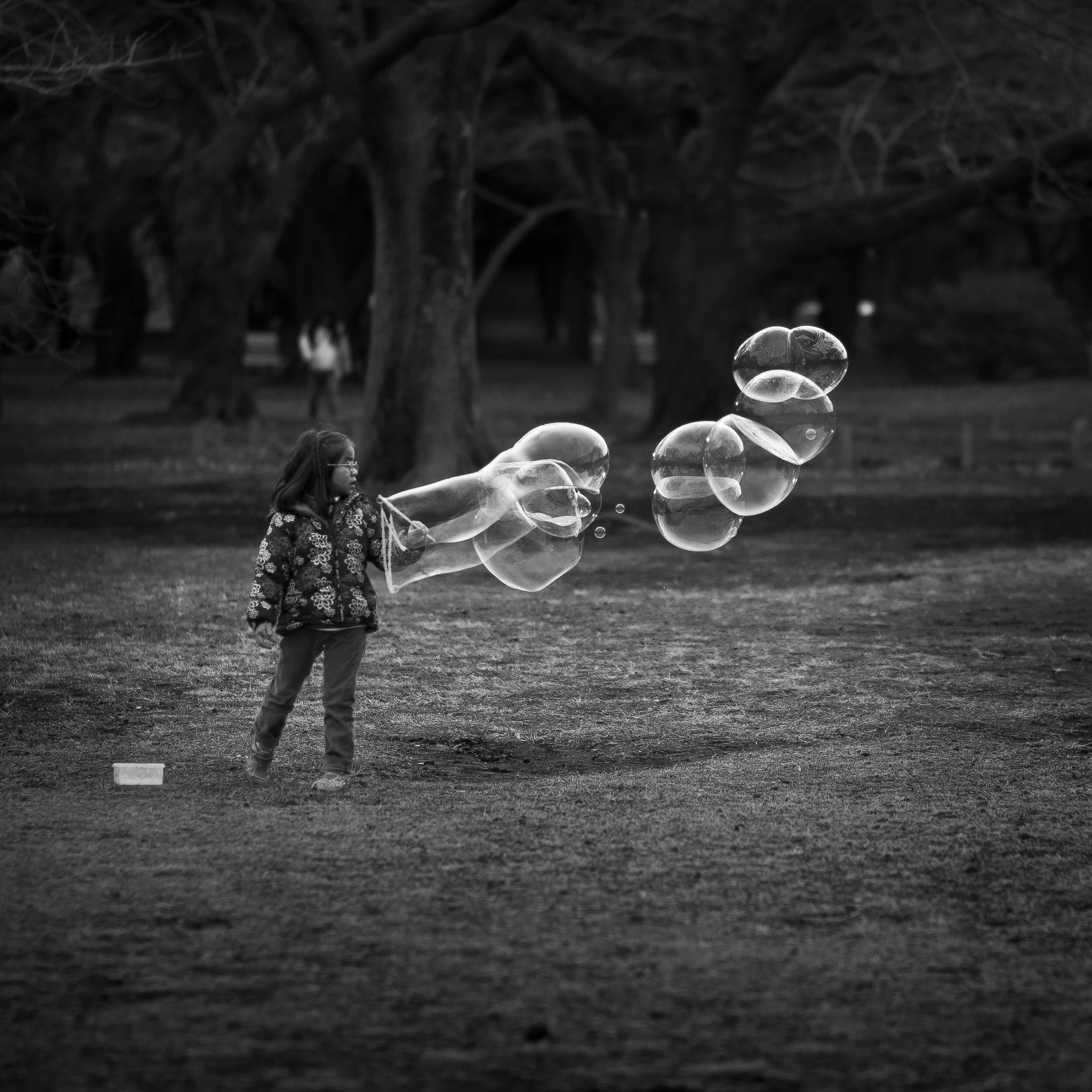 A young girl playing with soap bubbles in a park during evening or night, with trees and a blurred background.