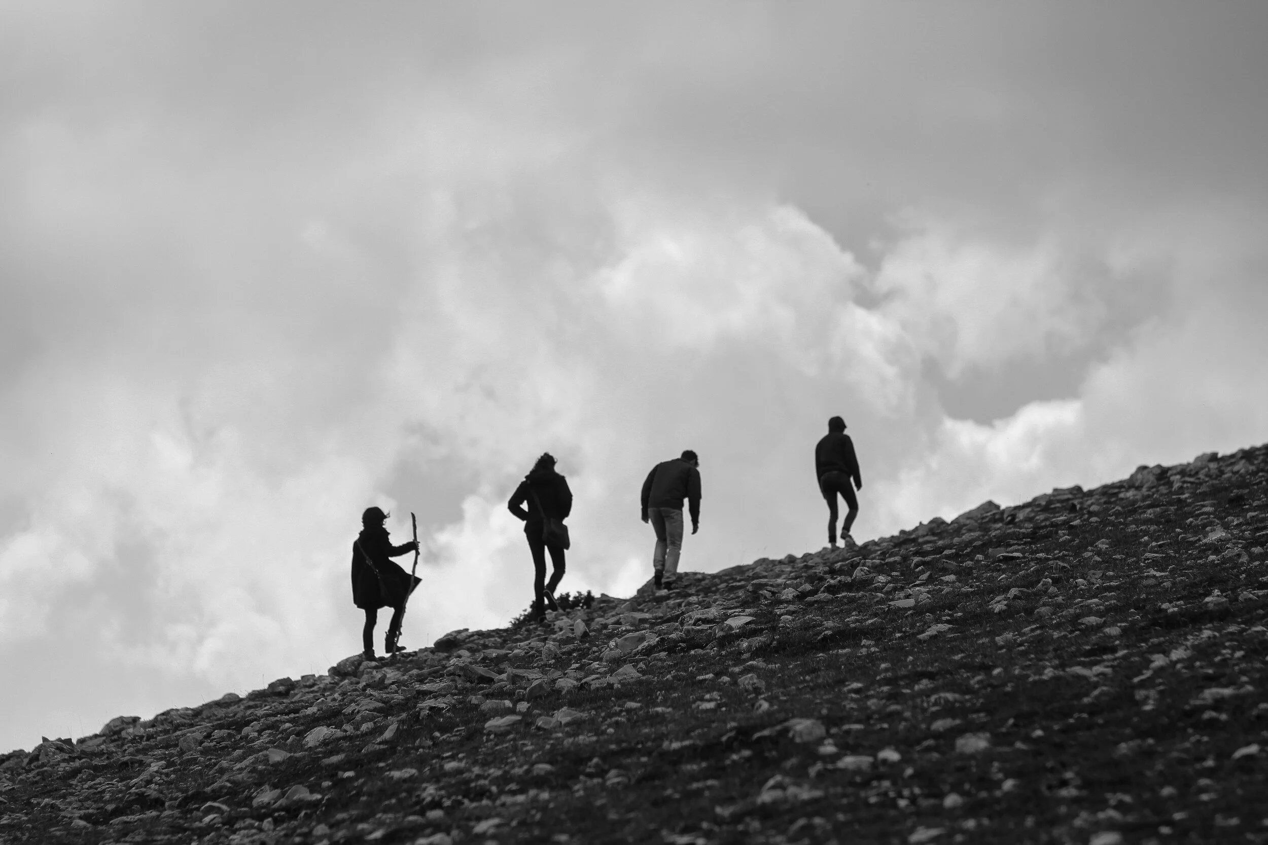 Silhouettes of five people hiking up a rocky slope against a cloudy sky.