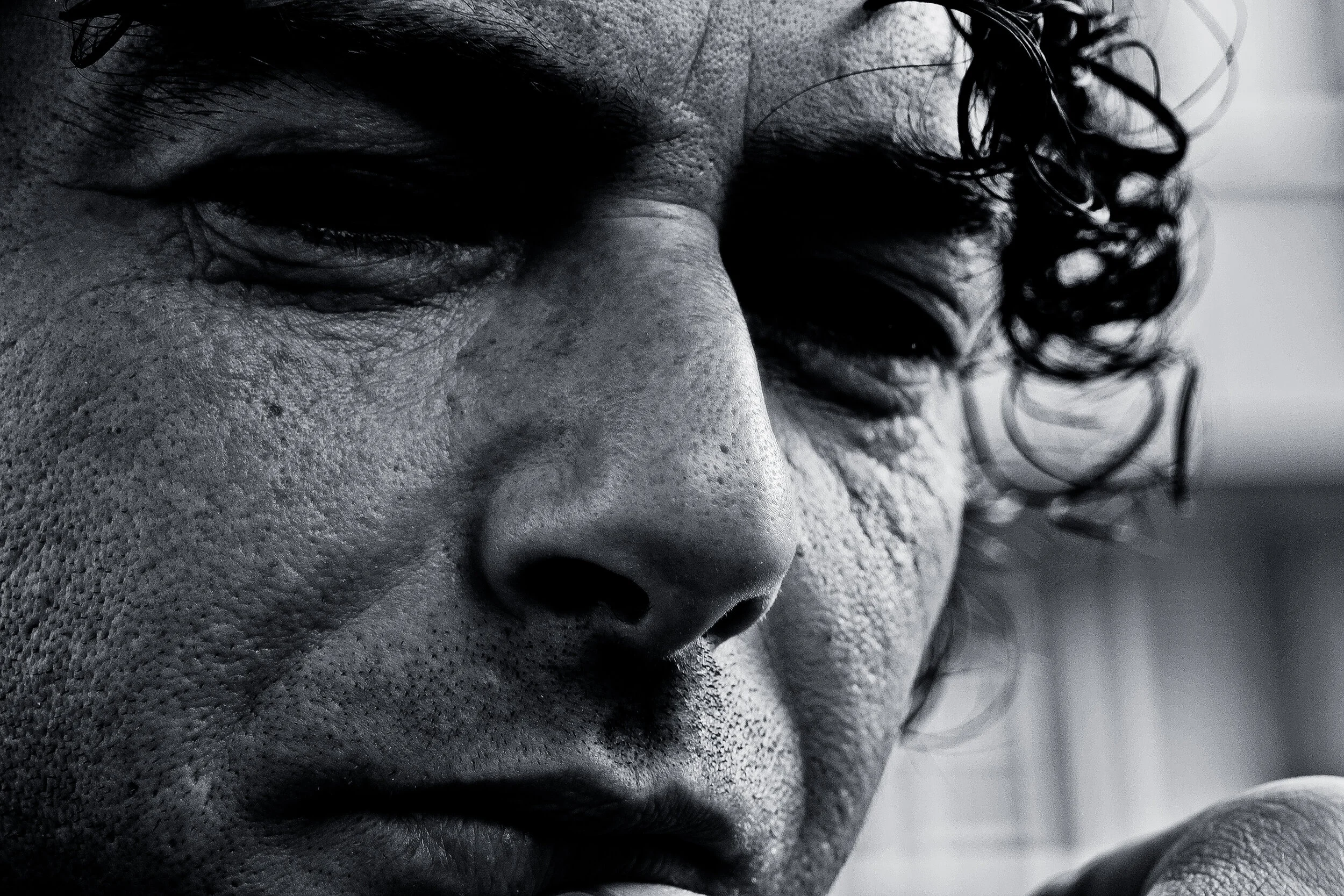 Close-up black-and-white photo of a person's face, focusing on the eyelid, nose, and part of the mouth, with curly hair partially covering the face.