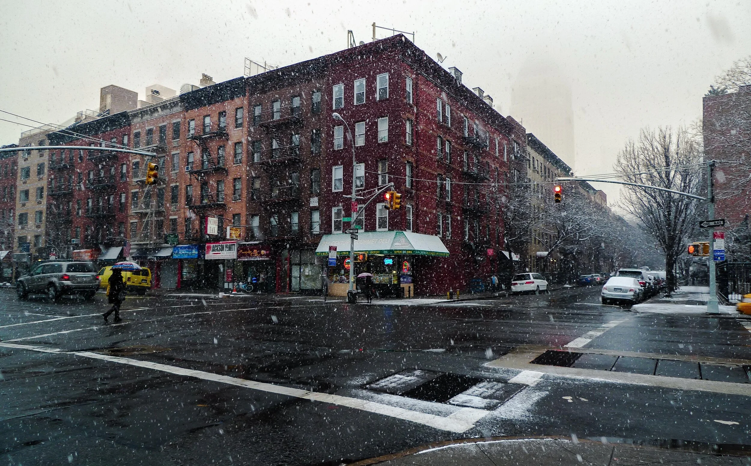 A city intersection on a rainy day with wet streets, cars, and pedestrians. A multi-story brick building with fire escapes and a storefront is visible, with snow falling in the scene.