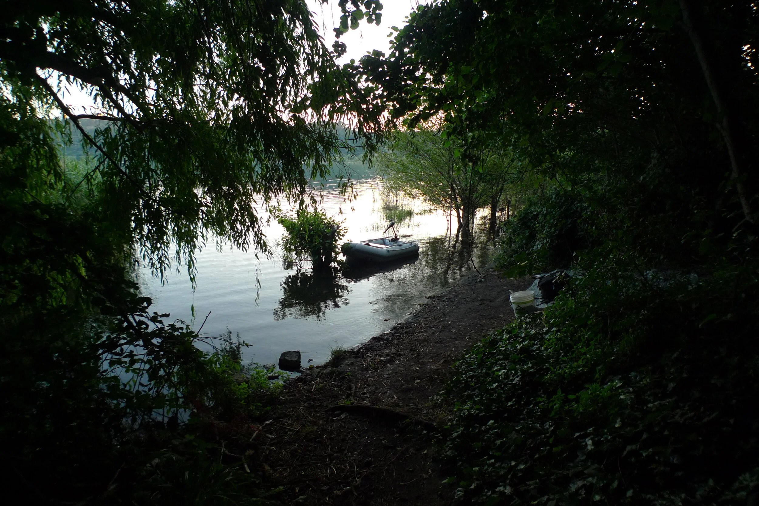 A secluded lakeside scene at sunset with dense green trees framing a small inflatable boat on the water and a bucket along the shore.