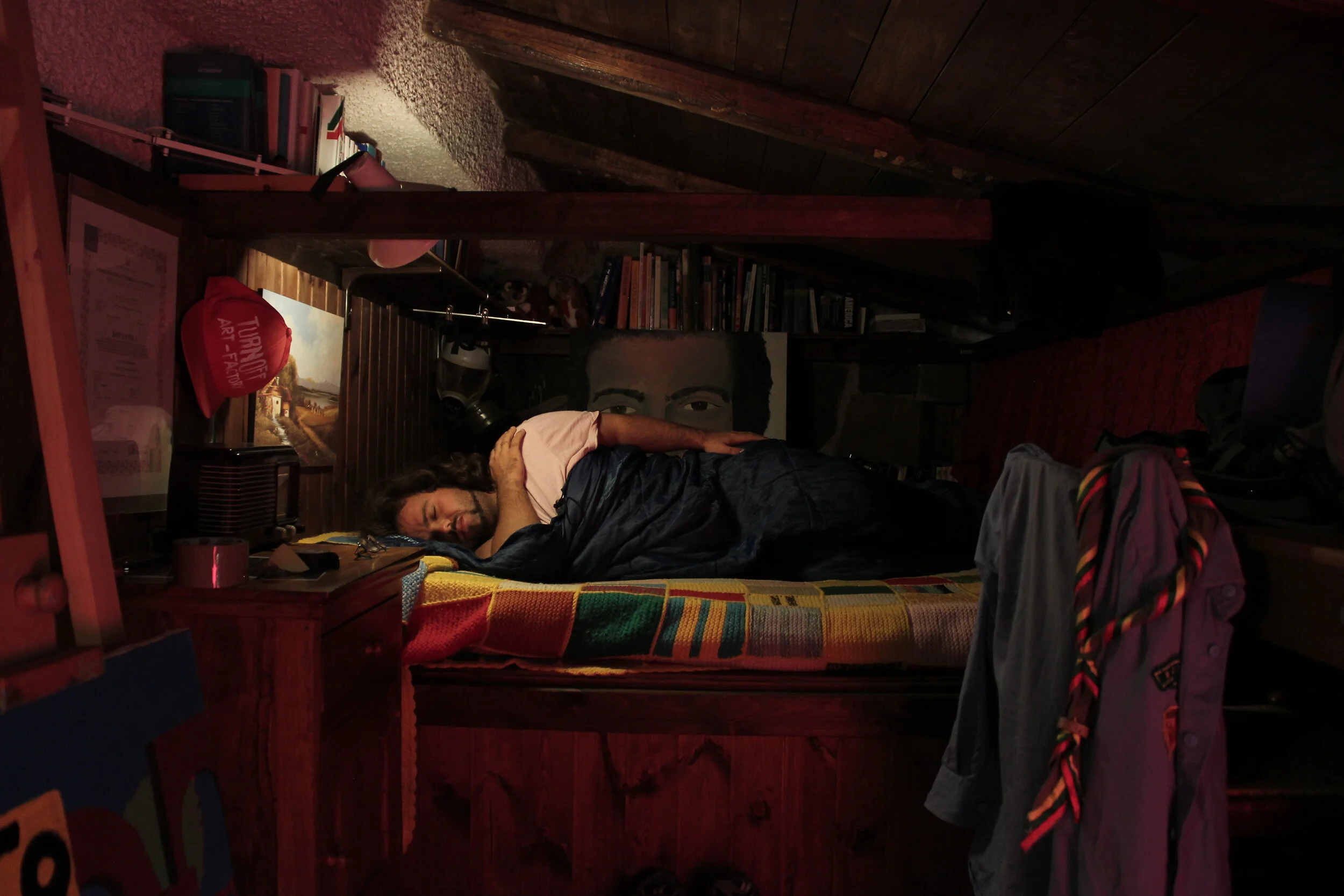A person with long hair lying on a bed covered with a colorful blanket in a cozy, dimly lit attic bedroom. The room has wooden walls, a bookshelf, a painting, and clothing hanging on the side.