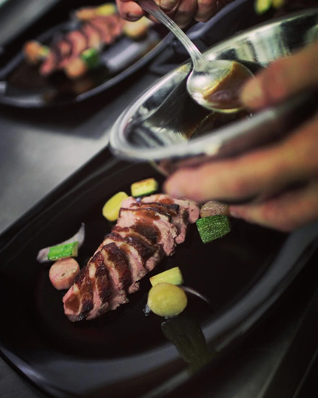 Pouring sauce over grilled meat with vegetables on a black serving dish.