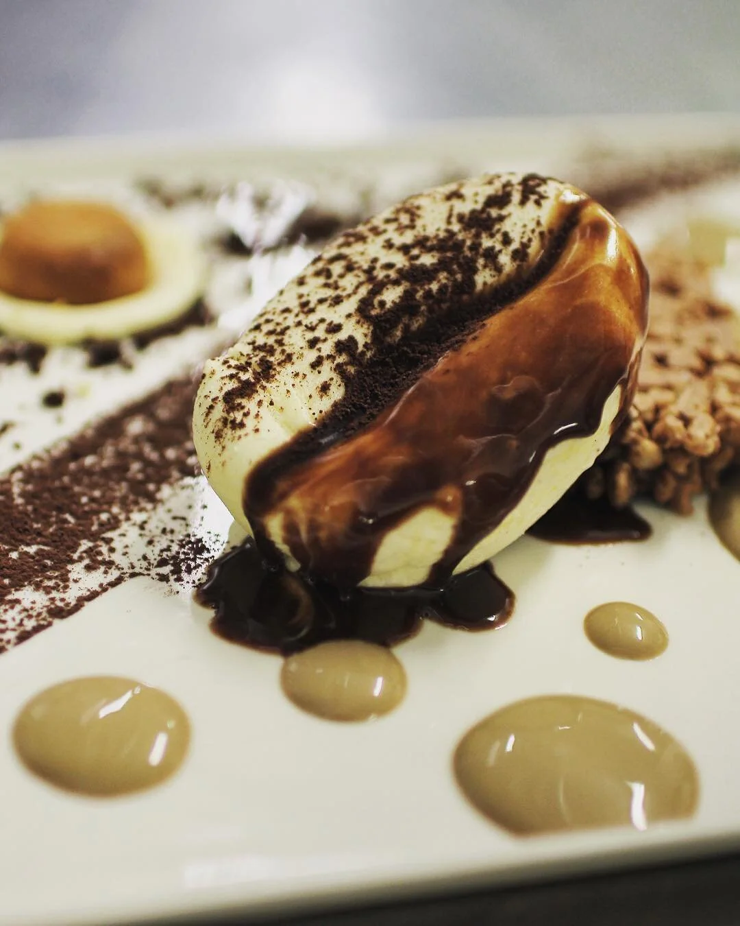 Close-up of a plated dessert with a scoop of ice cream drizzled with chocolate sauce, garnished with cocoa powder and small dollops of caramel sauce.