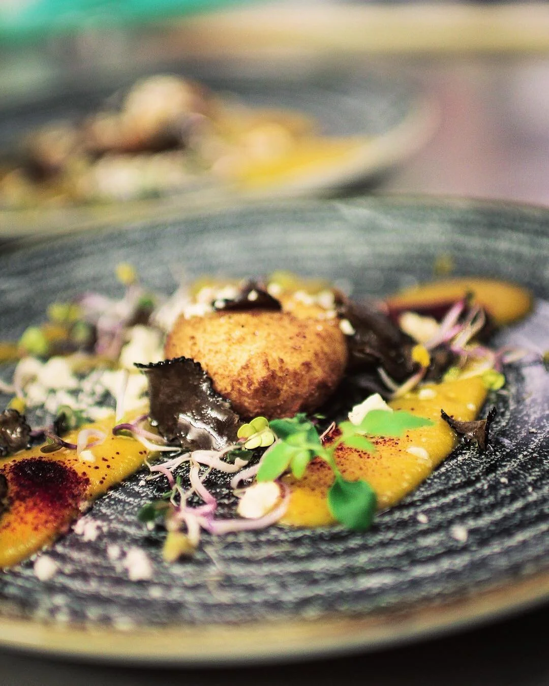 A gourmet dish featuring a fried ball surrounded by yellow sauce, garnished with microgreens, edible flowers, and dark chocolate shavings on a black textured plate.