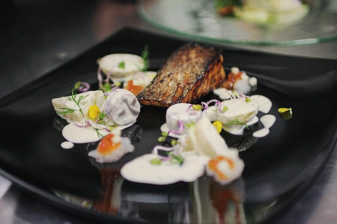 Grilled salmon fillet with sauce, garnished with microgreens and edible flowers, served on a black plate.