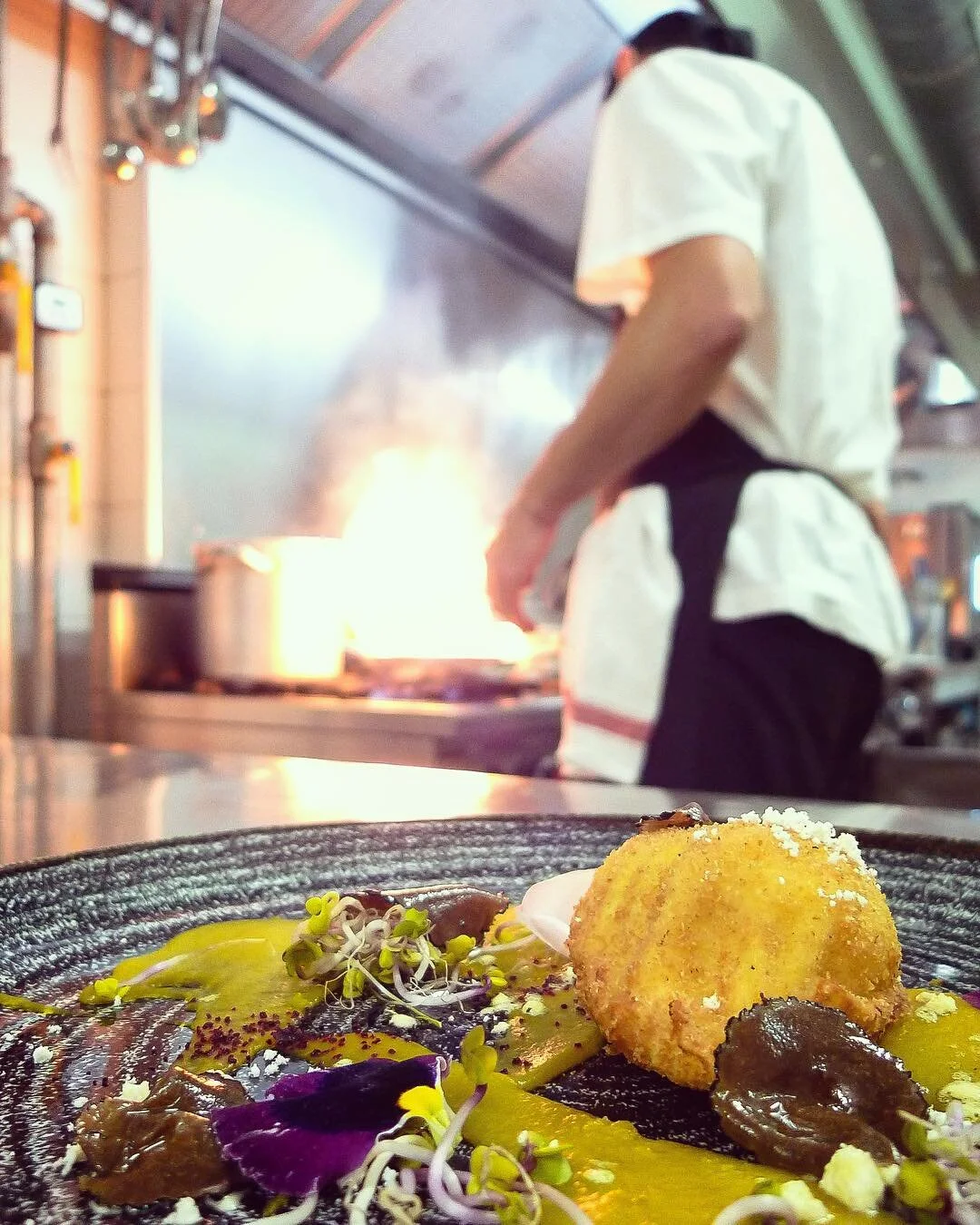 Close-up of a plated gourmet dish with a large, golden croquette, edible flowers, and various sauces, with a chef cooking in the background.