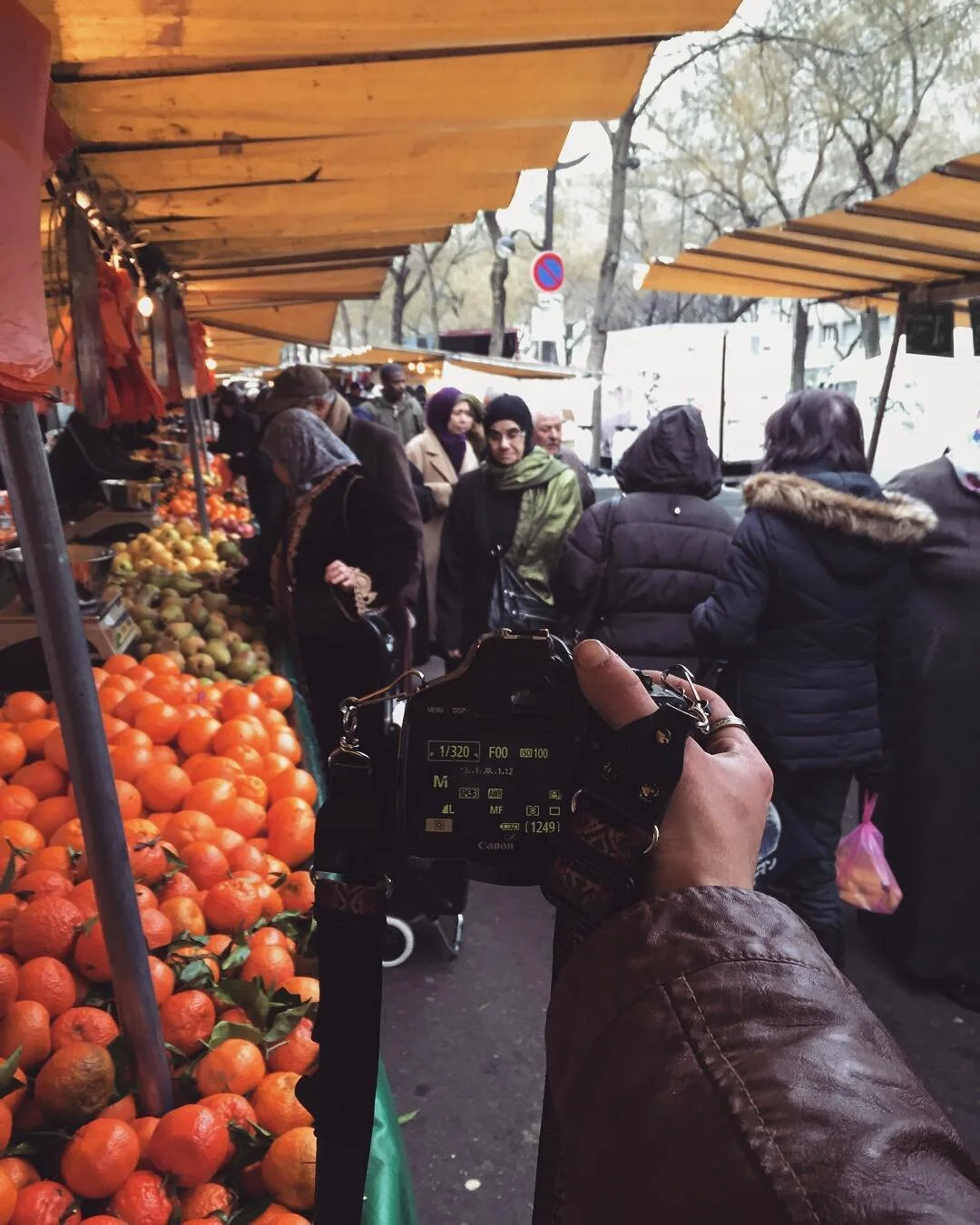 A person holding a camera at an outdoor market, with a produce stand filled with oranges and pears, and people shopping and browsing under yellow canopies in a busy outdoor market setting.