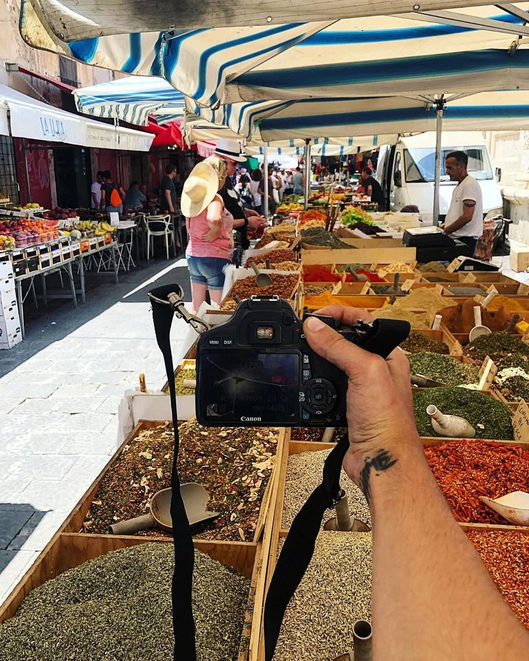 Person taking a photo at an outdoor marketplace with spice and herb stalls, vendors, shoppers, and striped umbrellas.