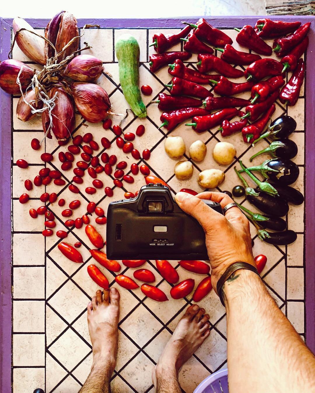A person taking a photo of various vegetables arranged on a tiled floor, including onions, a zucchini, chili peppers, cherry tomatoes, potatoes, and eggplants.