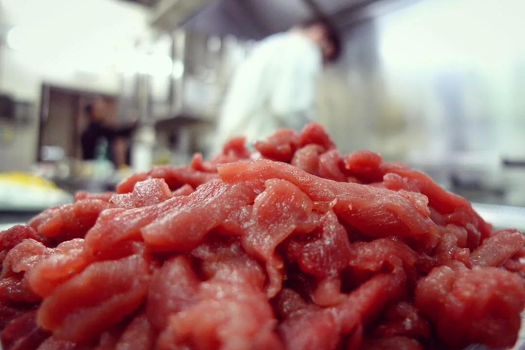 Close-up of raw, diced meat on a plate in a kitchen, with a blurred background including a person wearing a white coat.