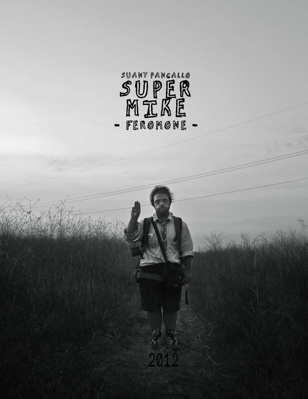 A black and white photo of a man walking down a trail through tall grass, waving at the camera, with a cloudy sky above. Text overlay includes "Suany Pangalelo," "Super Mike," "Feromone," and "2012."