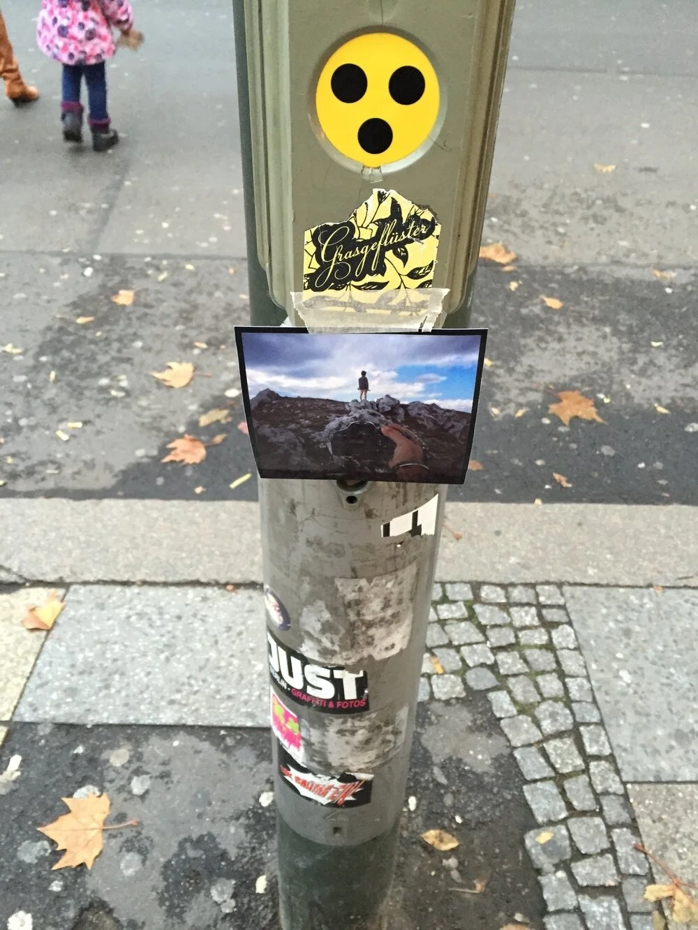 A metal pole with various stickers and photos attached, including a yellow circular sticker with three black dots resembling a surprised face, a sticker with the word 'Grasgelfleister,' and a smaller photo of a person standing on a rocky terrain look