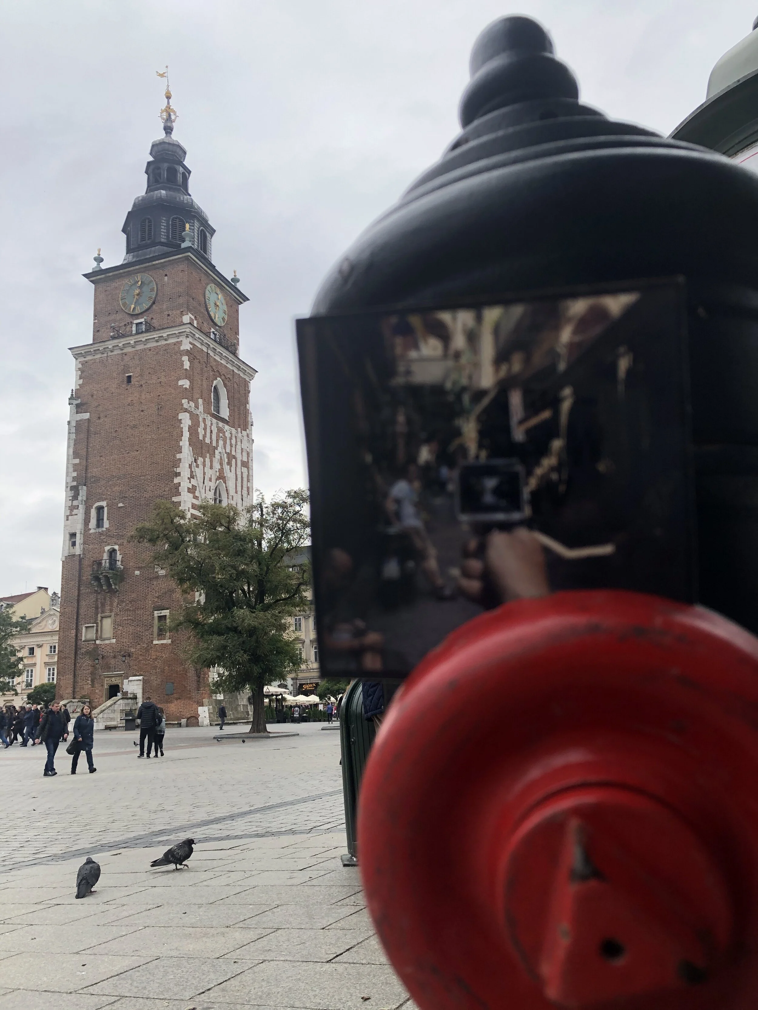 A clock tower with a brick facade and a large clock face, seen behind a large red fire hydrant in a city square with people walking and pigeons on the ground.