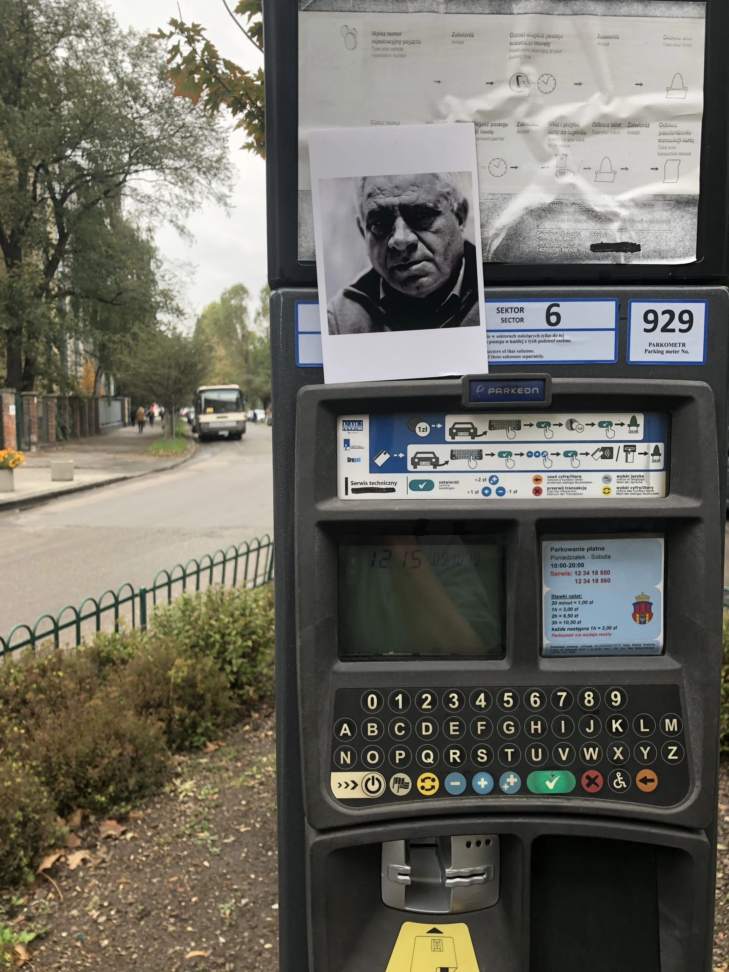 Parking meter with a black and white photo of a man's face pinned to it. The parking meter shows the time 12:15, with various instructions and symbols in Polish. The surrounding area features a sidewalk, some trees with green leaves, and a city stree