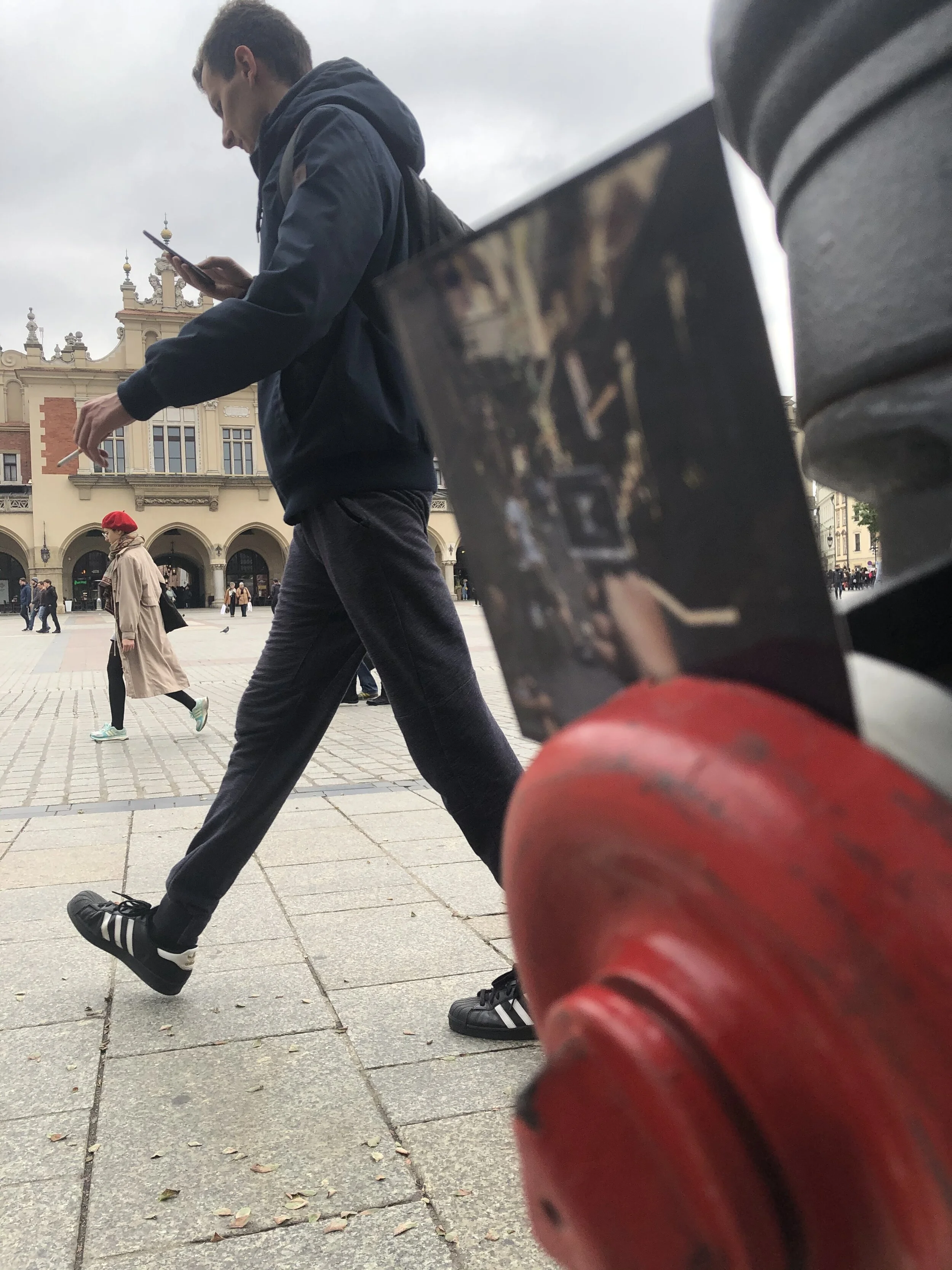 A young man with a backpack walking on a city square, looking at his phone, with a woman in a red beret walking behind him, and a historic building in the background. There's a blurred red structure in the foreground.