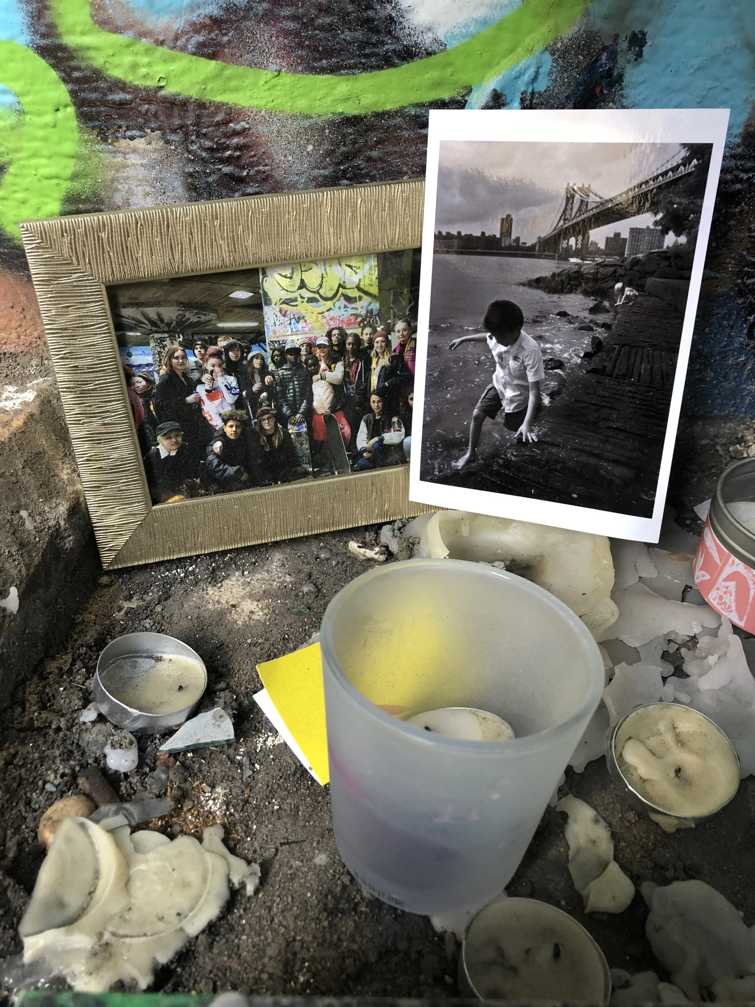 A collection of used candles, some in containers and some melted and spilled, surrounded by debris on a dirt surface. A framed photo of a group of people is leaning against a surface, and a black and white photo of a boy by a river with a bridge is p