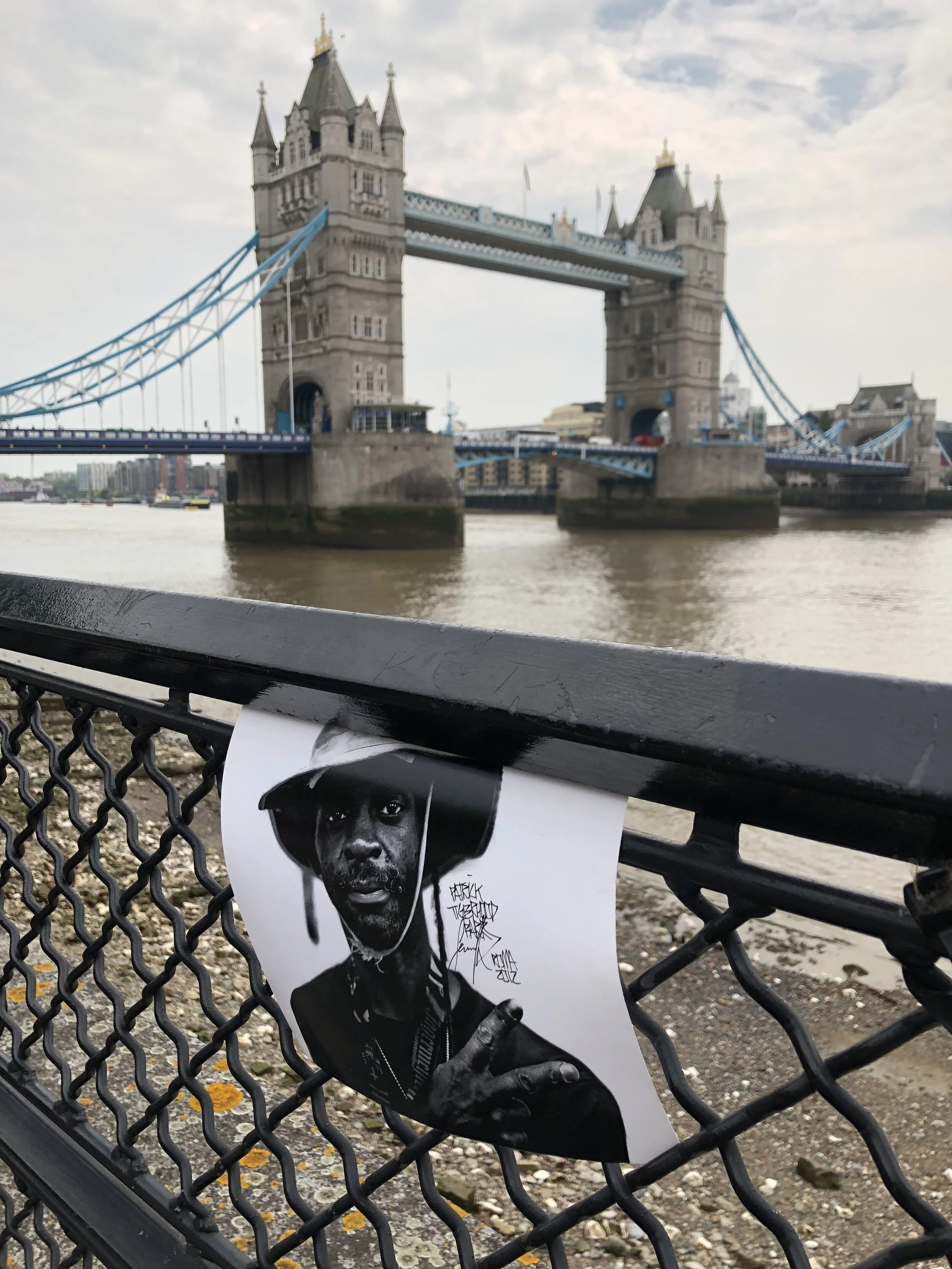 Photo of Tower Bridge in London, seen from across the River Thames, with a black and white photograph of a man wearing a hat and holding a cigarette attached to a fence in the foreground.