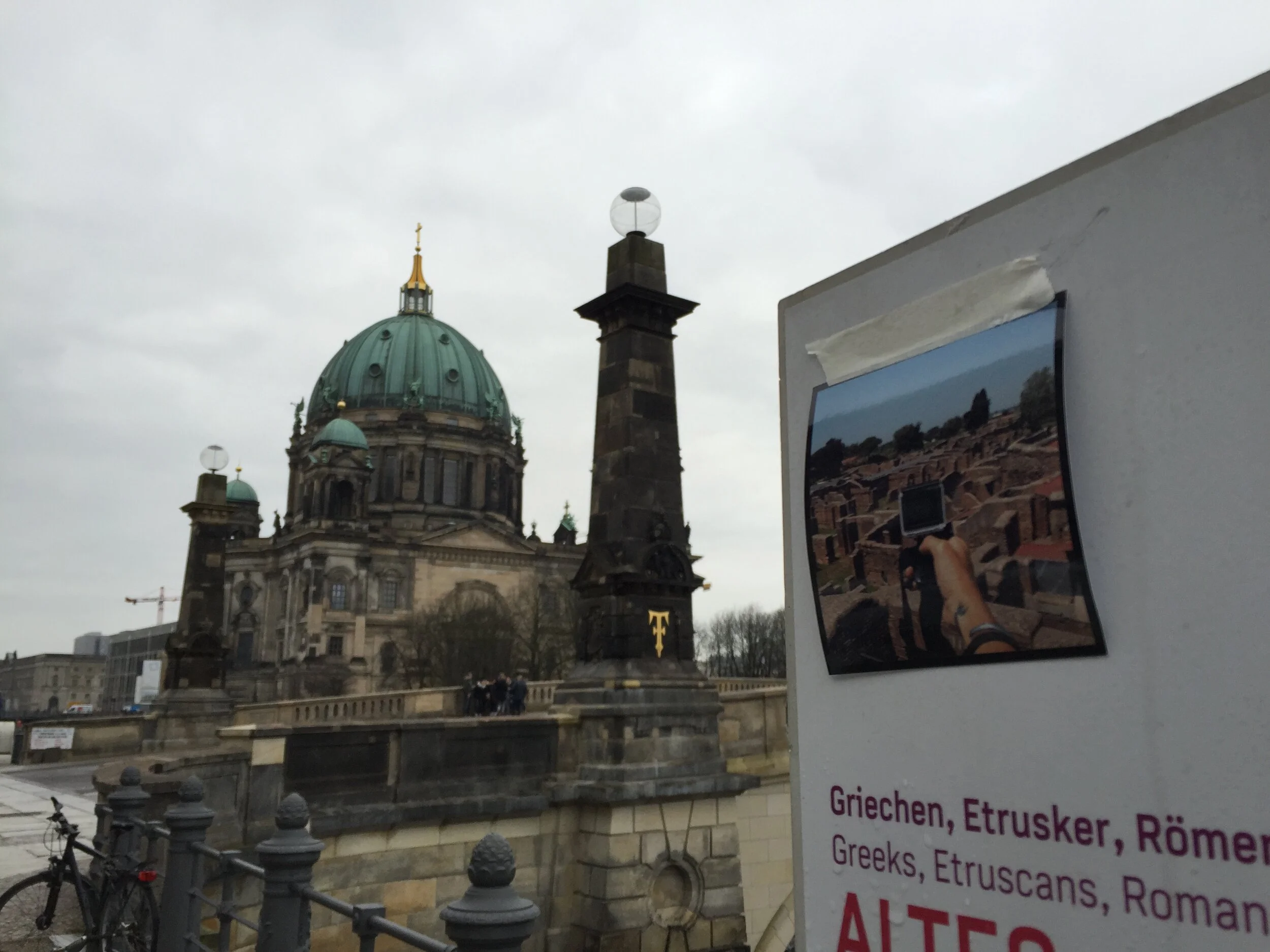 View of a historic European cathedral with a large green dome, ornate details, and a smaller domed tower in front of a cloudy sky. Part of a white poster with partially visible text and a bicycle in the foreground.