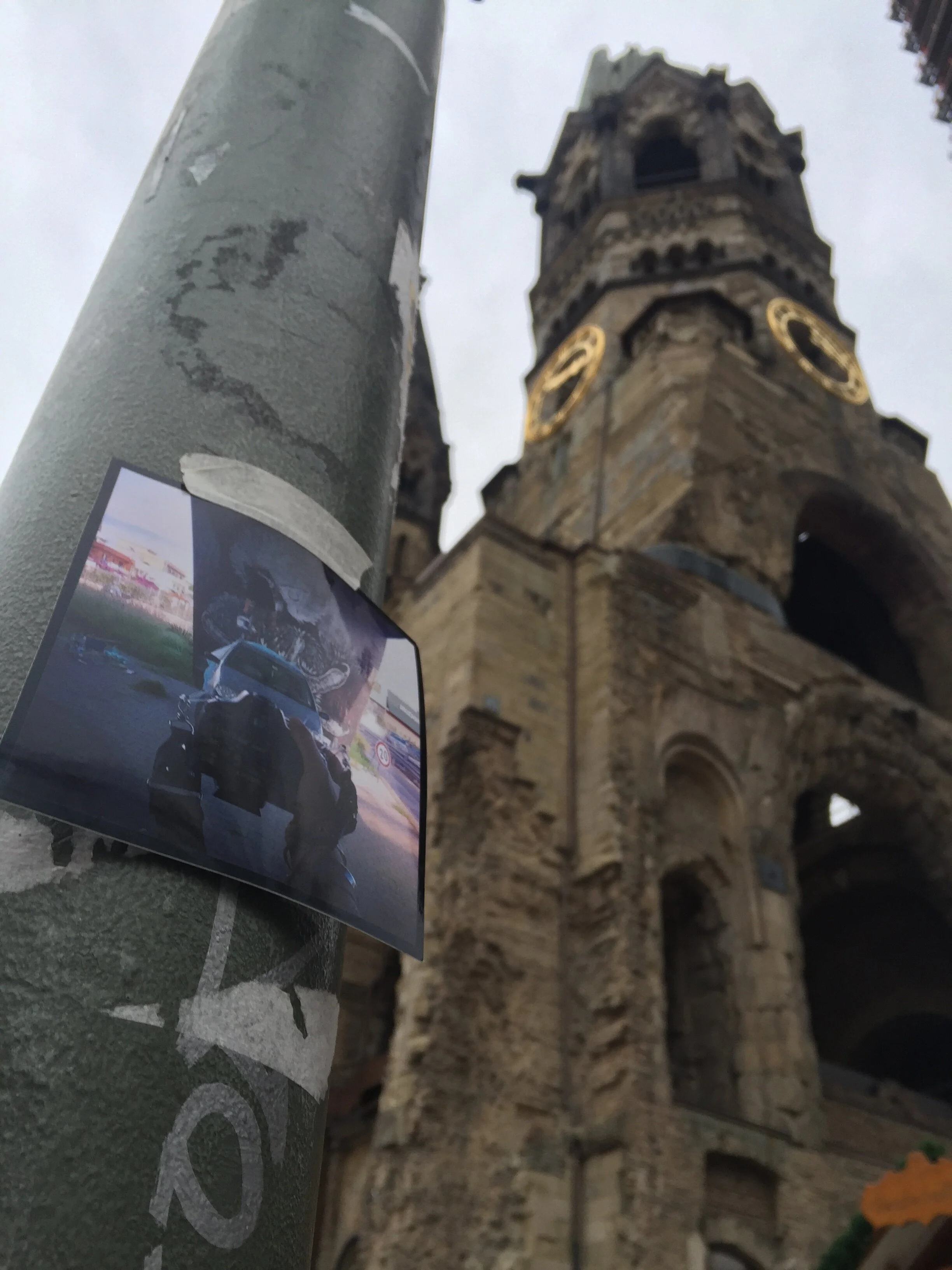 Close-up of a sticker on a pole with a historic stone building with clock faces in the background, under an overcast sky.