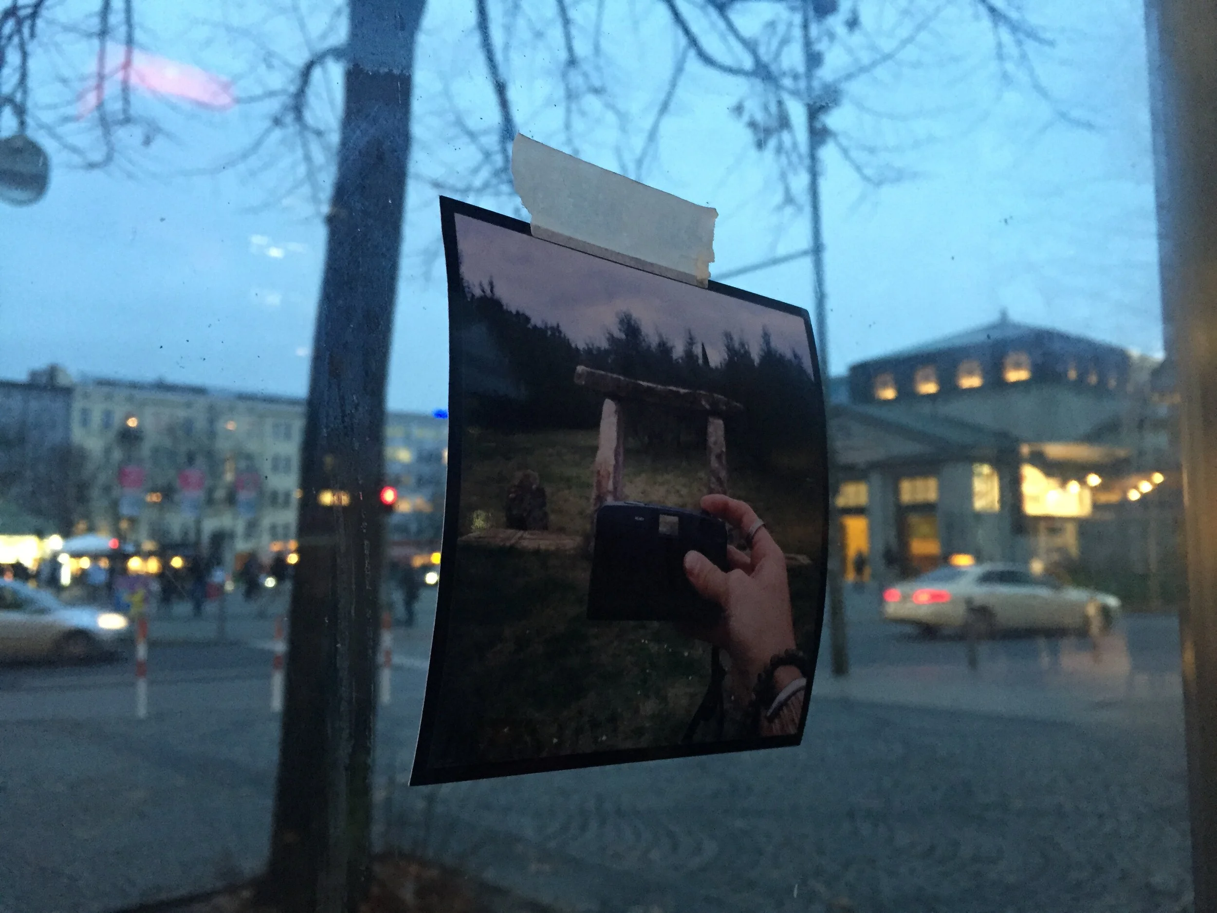 A window looking out onto a cityscape with buildings illuminated at dusk or dawn, leafless trees are visible outside, and a reflection of a hand holding a phone capturing the image is visible on the glass.