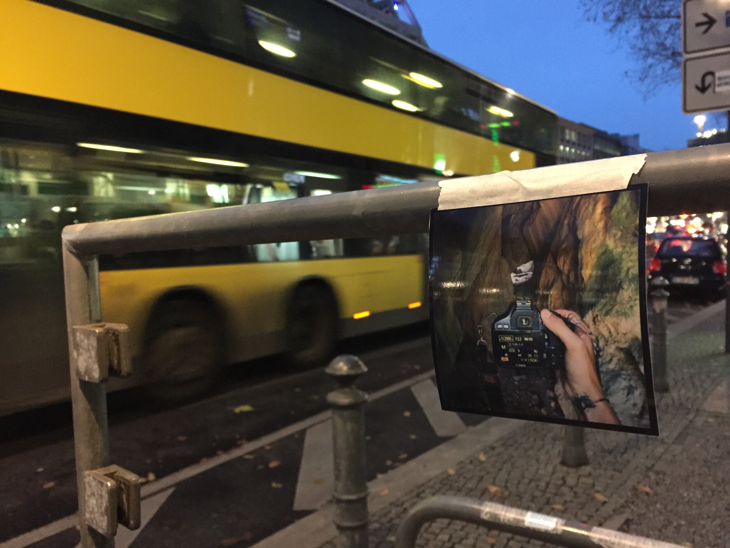 A person holding a camera is reflected in a painting of a landscape, which is propped against a railing at an outdoor bus stop during nighttime. In the background, a yellow bus is passing by, and there are city buildings with lit windows. There are a