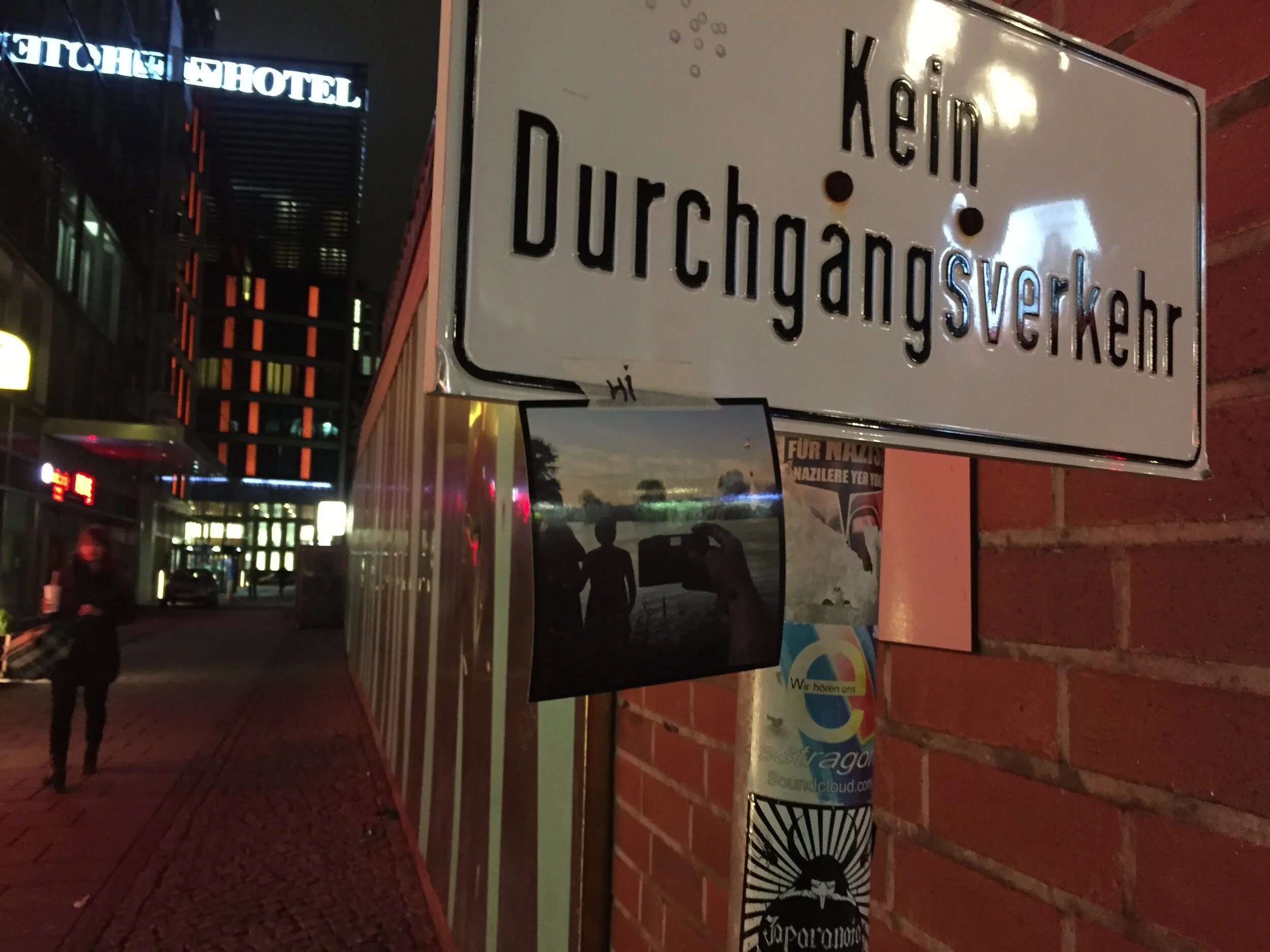 Street scene at night with a sign in English and German, and a building with illuminated signage in the background.