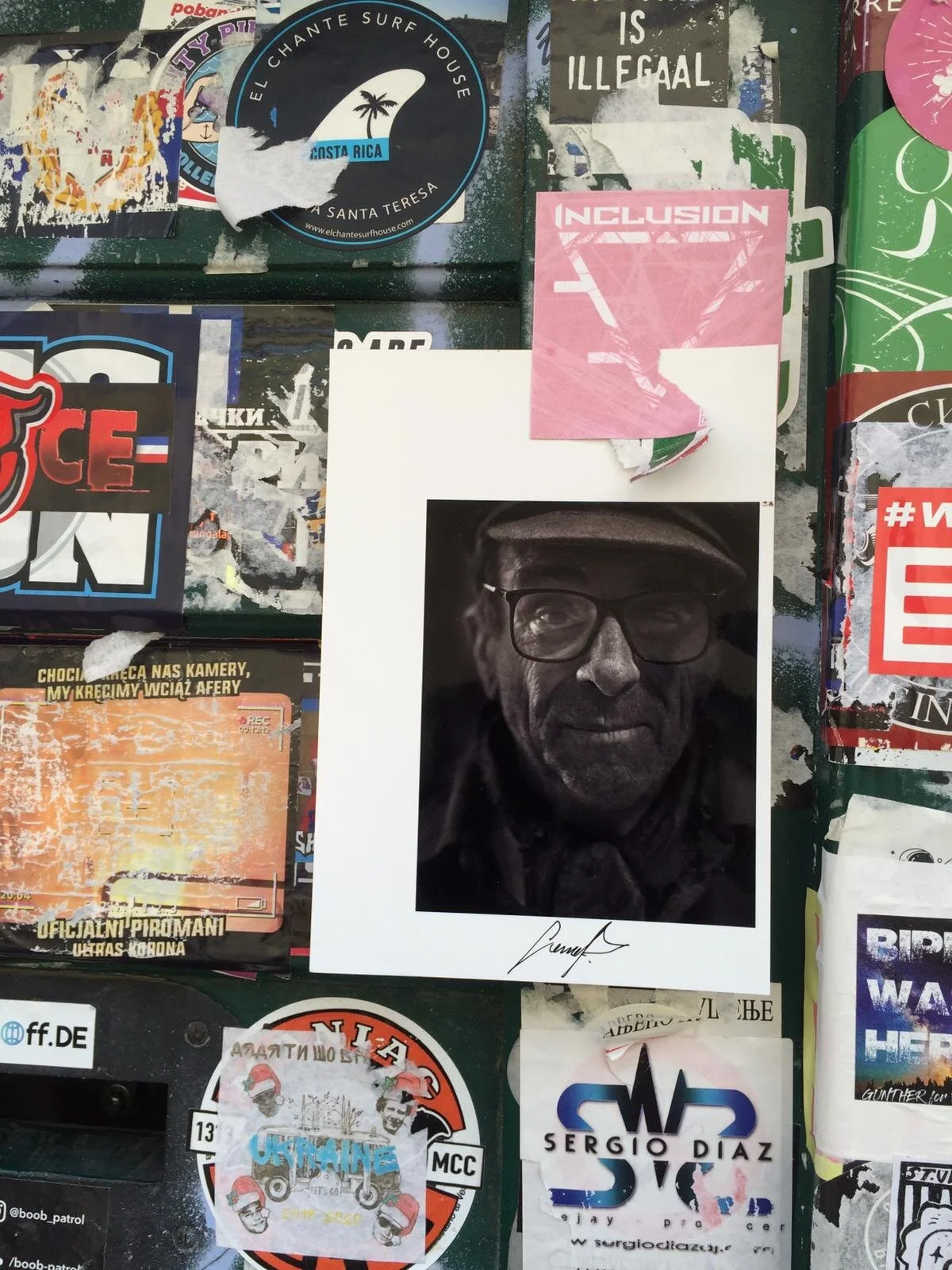 A collage of various stickers and posters on a public surface. Prominent items include a black-and-white portrait of an older man wearing glasses and a cap, signed at the bottom; a pink sticker with the word 'INCLUSION'; a round sticker with a surfbo