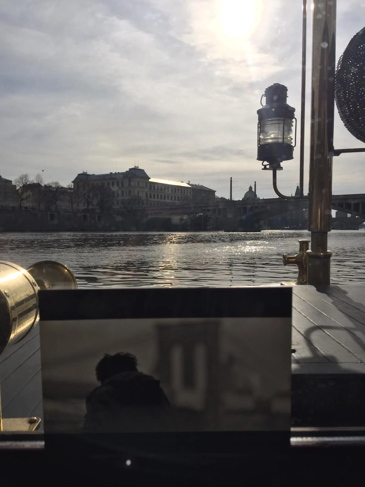 View of a river with buildings and a bridge in the distance, taken from a boat with a lantern and railing in the foreground, under a cloudy sky with sunlight.