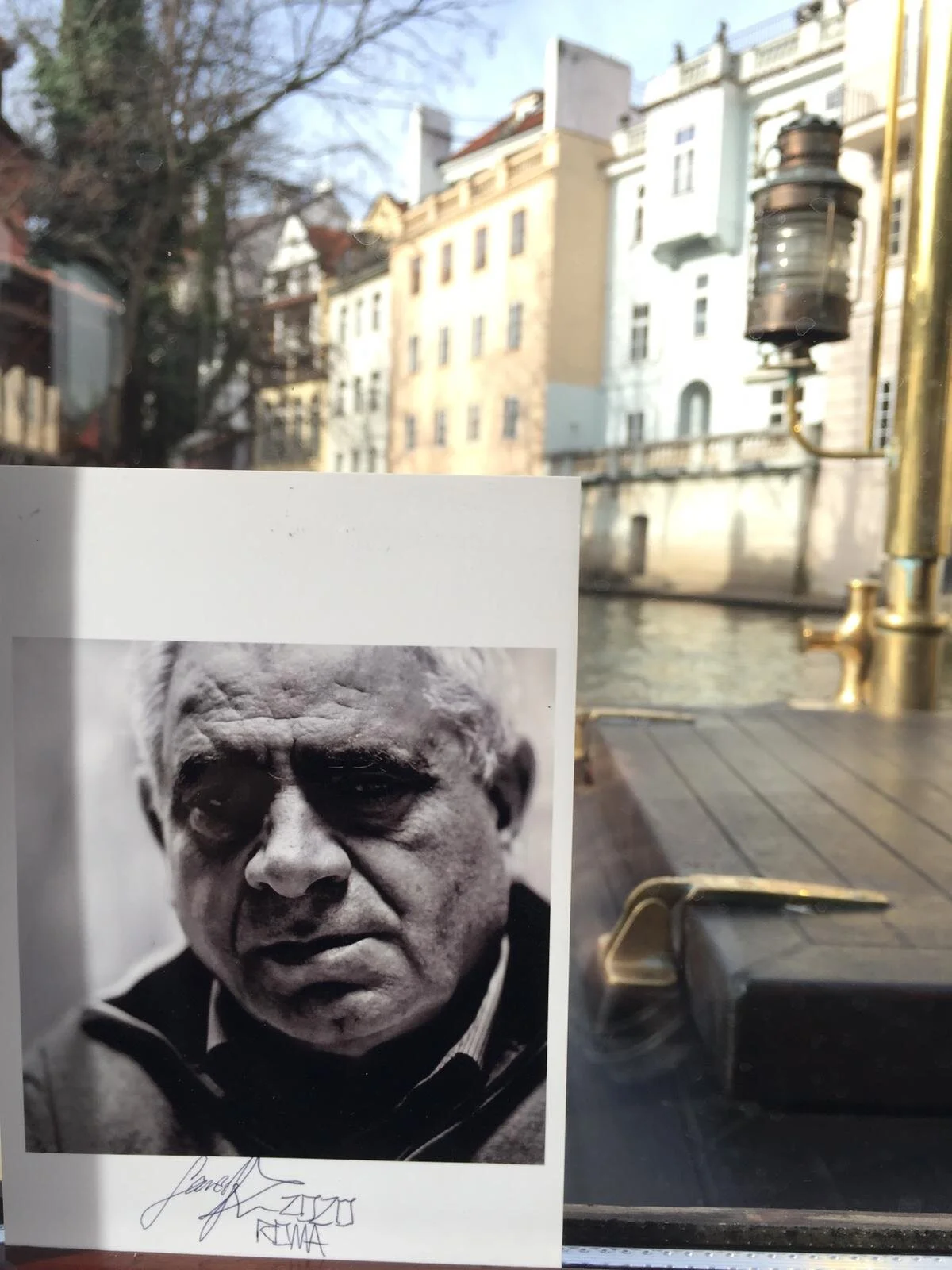 A black-and-white photograph of an elderly man with a serious expression, signed at the bottom, held up against a window showing buildings, trees, and a canal outside.