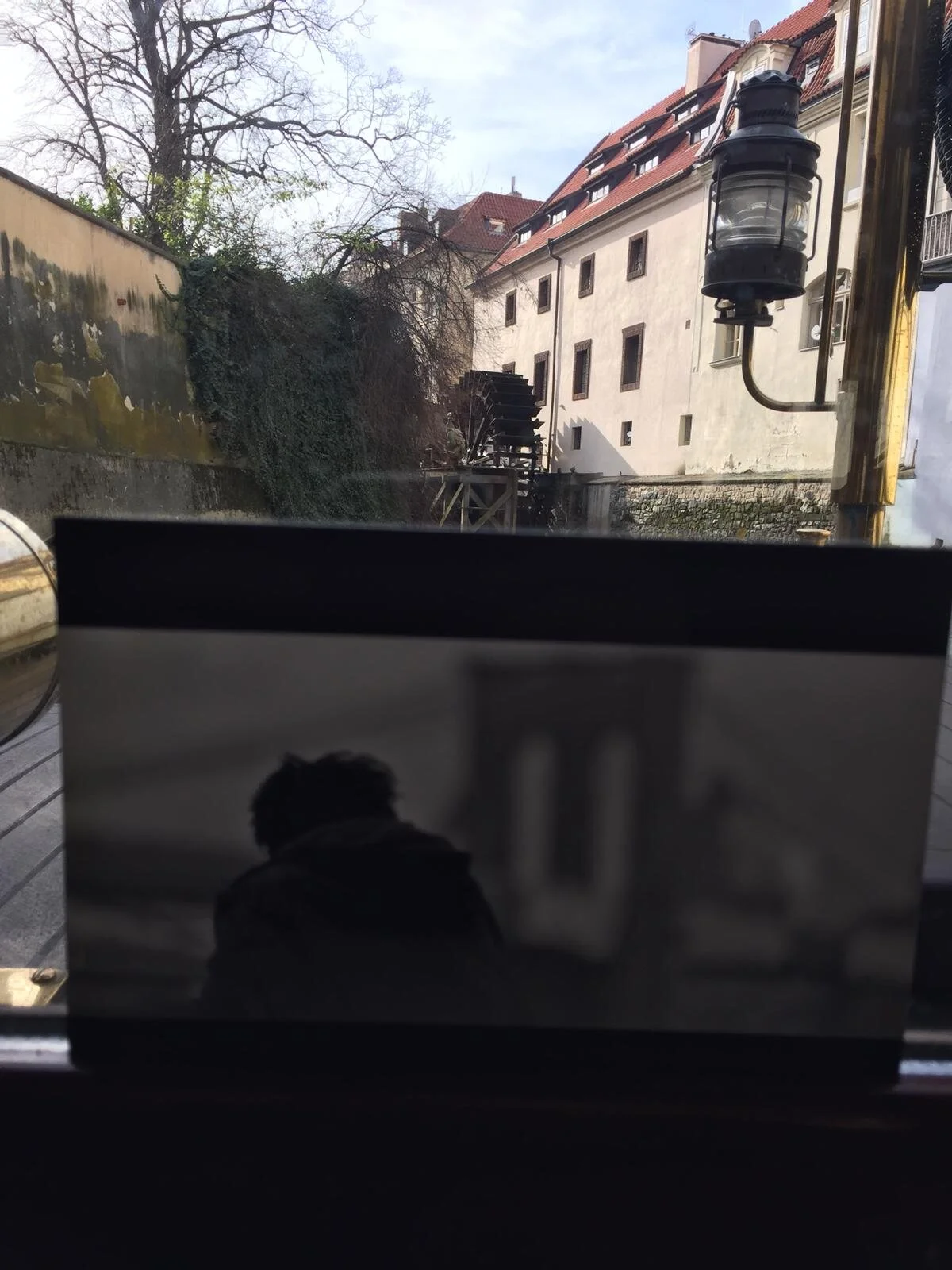 Looking through a window with a reflection of a person taking a photo indoors, showing a building with a water wheel and staircase outside.