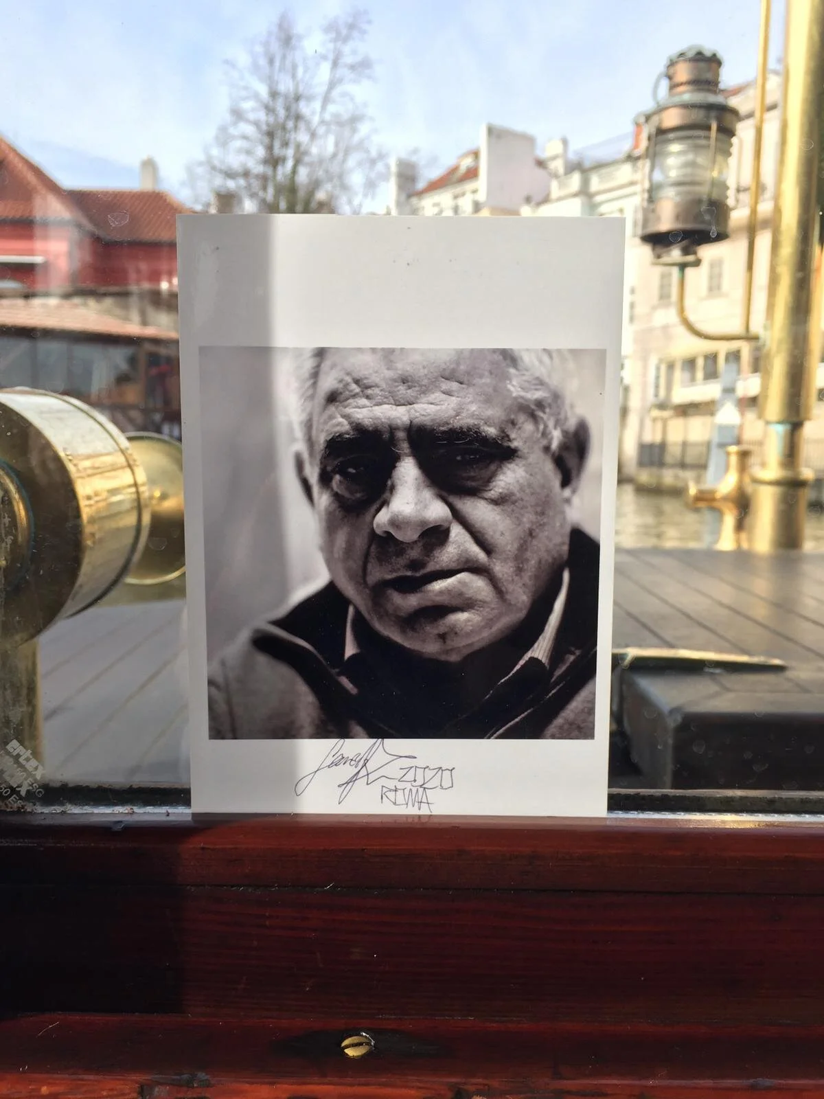 Black and white photograph of a serious-looking older man with short gray hair, signed below the photo, displayed on a window with a cityscape in the background.