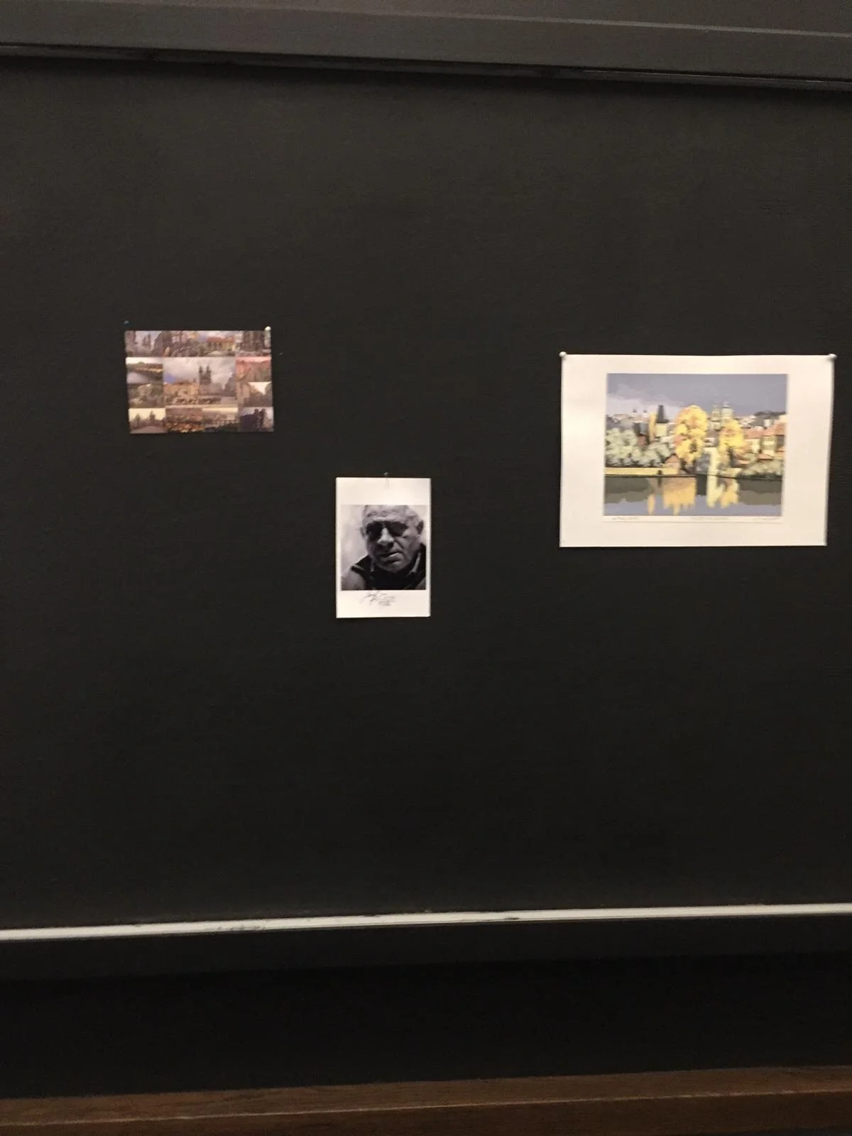 A black wall with three photographs displayed. The left photo is a collage of multiple images, the right photo is a landscape painting or print of a cityscape with trees reflected in water, and the center photo is a black-and-white portrait of an eld