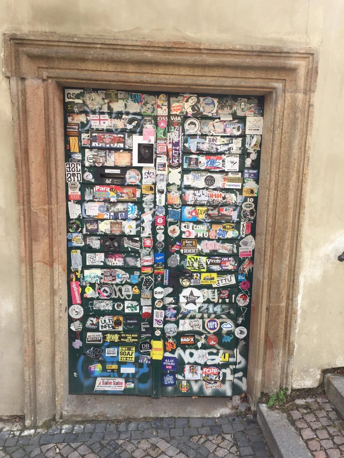 A hand-painted double door covered in a large collection of stickers and decals, with a stone frame around it, located outdoors on a stone pavement.