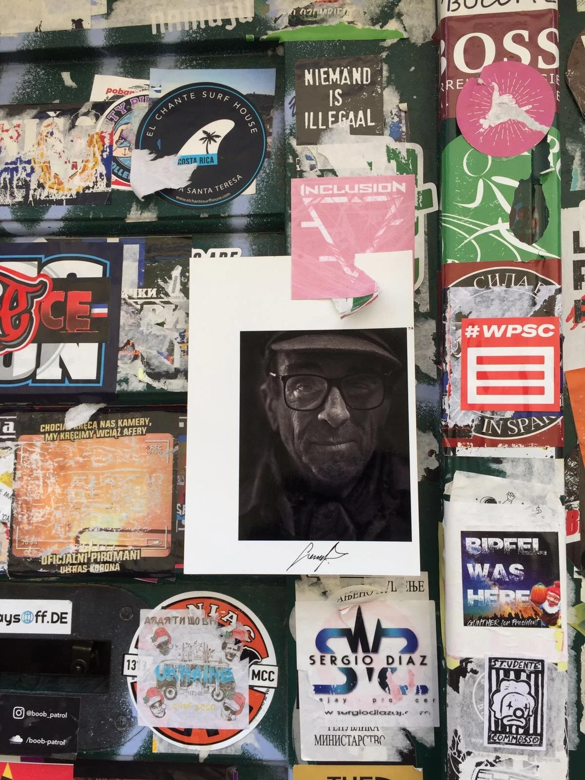 A collage of various stickers, posters, and flyers on a surface, including a black and white photo of an older man wearing glasses and a hat, with a signature below, and several other colorful stickers and signs with text in different languages.